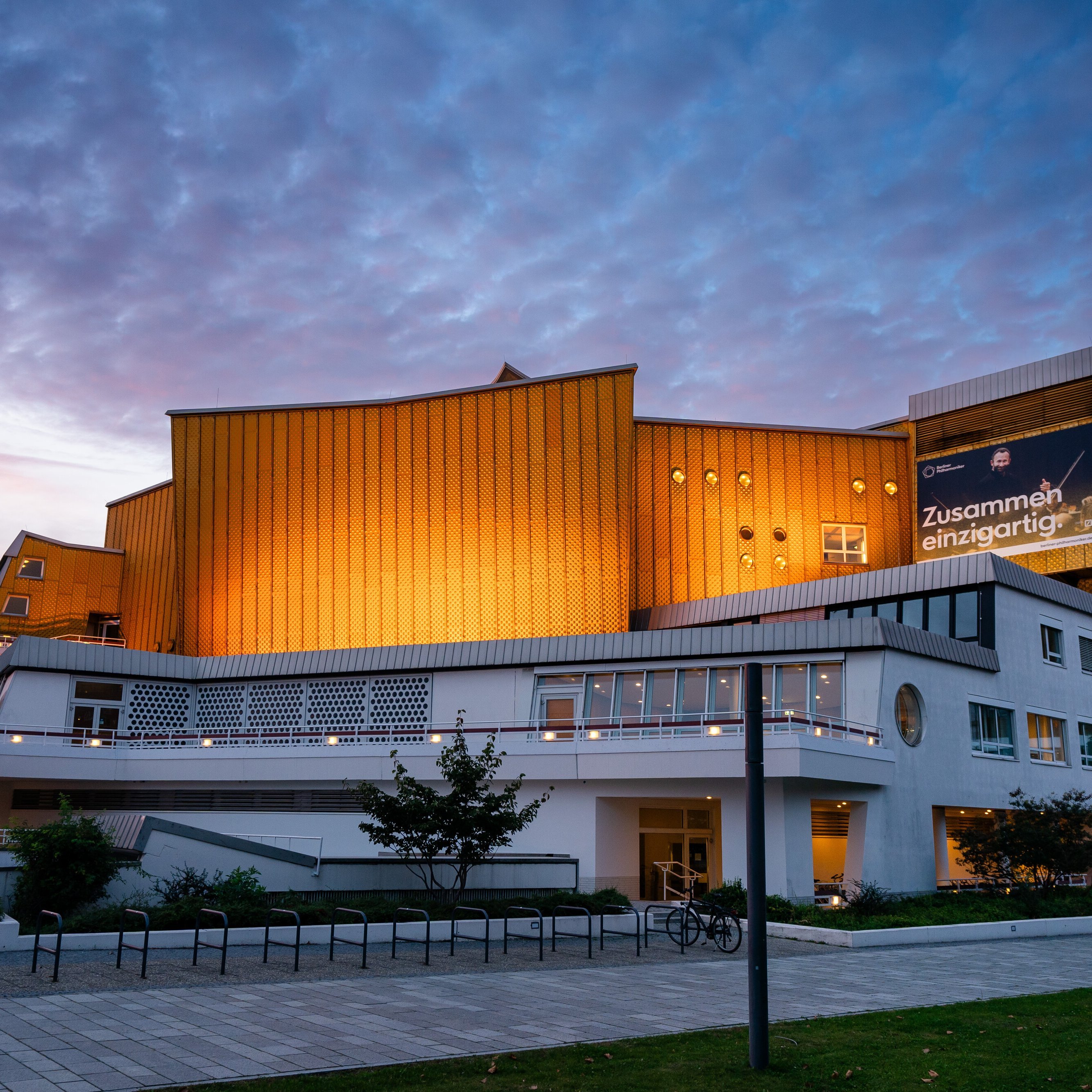 Die Philharmonie Berlin mit goldenen Paneelen und weißen unteren Stockwerken wird in der Abenddämmerung beleuchtet, mit einem bewölkten Himmel im Hintergrund. Auf einem großen Banner steht auf Deutsch »Zusammen einzigartig!.