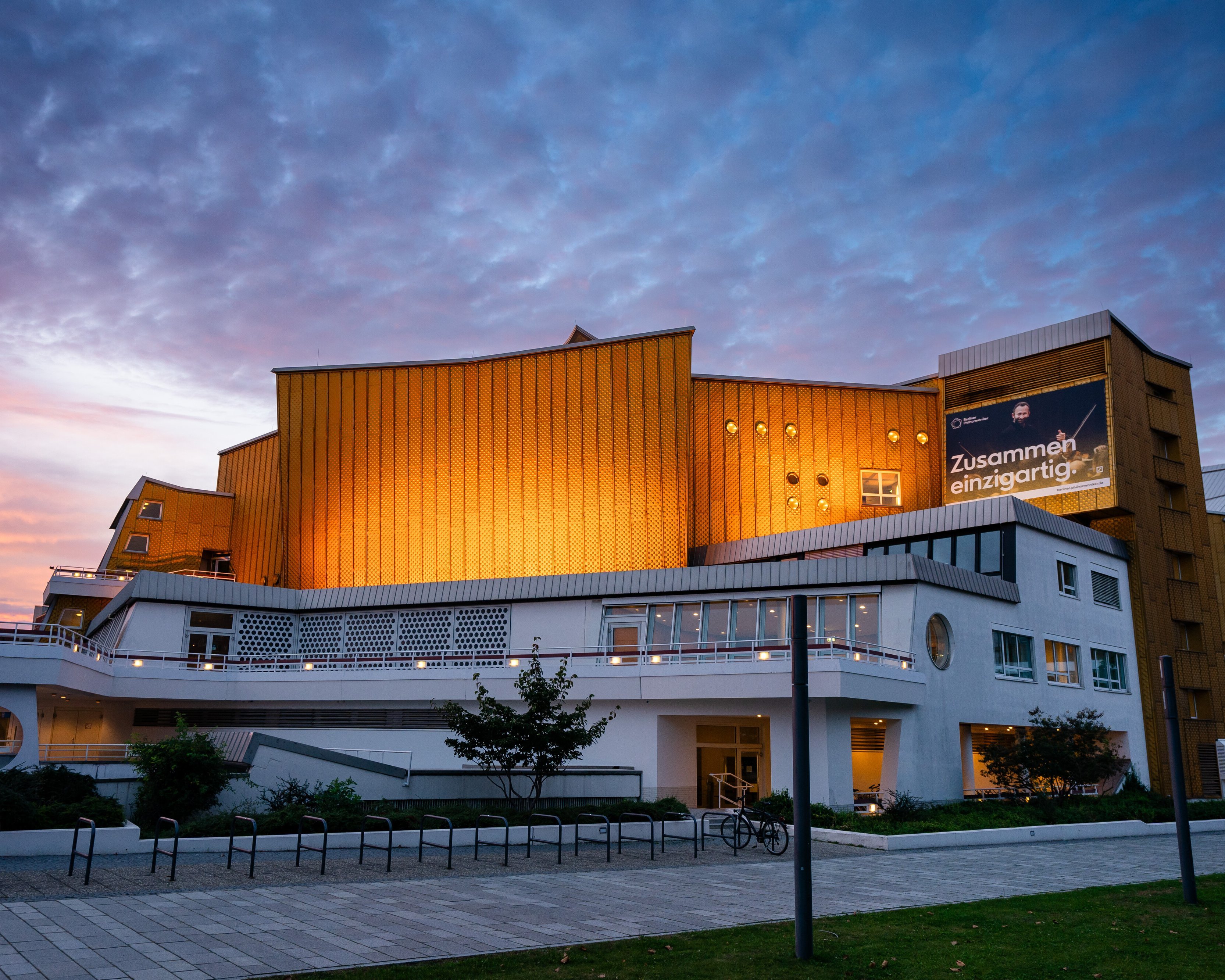 Die Philharmonie Berlin mit goldenen Paneelen und weißen unteren Stockwerken wird in der Abenddämmerung beleuchtet, mit einem bewölkten Himmel im Hintergrund. Auf einem großen Banner steht auf Deutsch »Zusammen einzigartig!.
