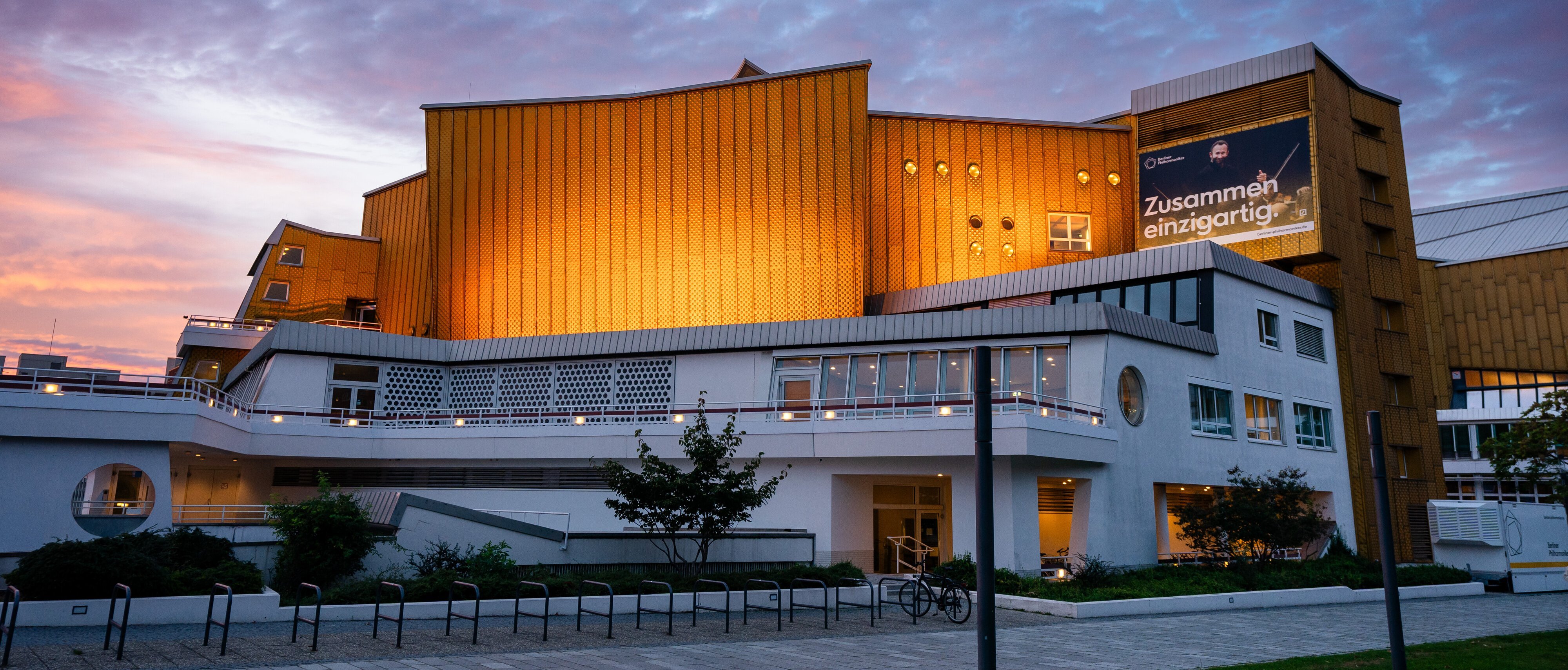 Die Philharmonie Berlin mit goldenen Paneelen und weißen unteren Stockwerken wird in der Abenddämmerung beleuchtet, mit einem bewölkten Himmel im Hintergrund. Auf einem großen Banner steht auf Deutsch »Zusammen einzigartig!.