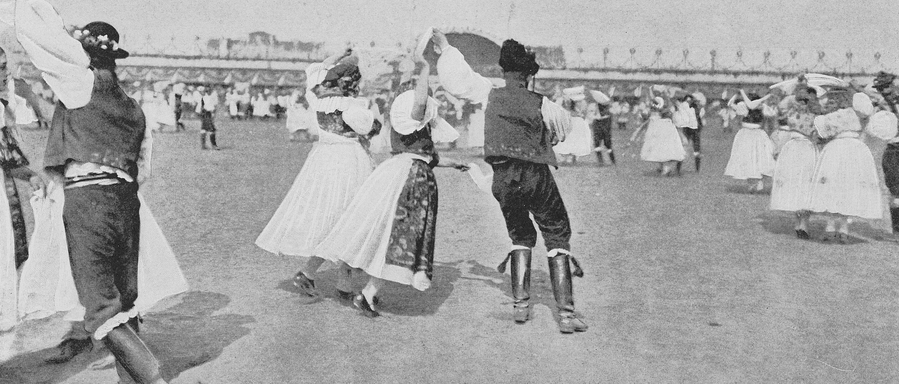 Männer und Frauen in traditionellen Trachten tanzen paarweise auf einer Wiese, während andere Gruppen im Hintergrund bei hellem Tageslicht tanzen.