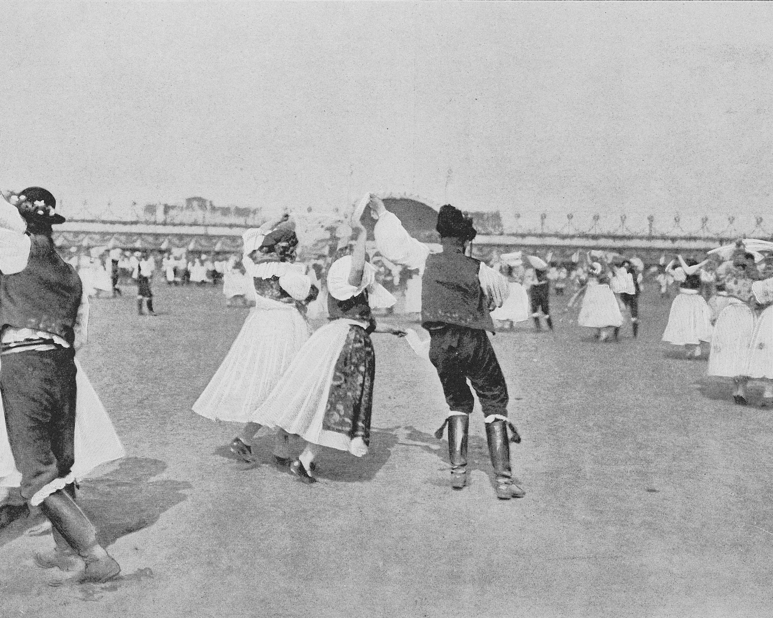 Männer und Frauen in traditionellen Trachten tanzen paarweise auf einer Wiese, während andere Gruppen im Hintergrund bei hellem Tageslicht tanzen.
