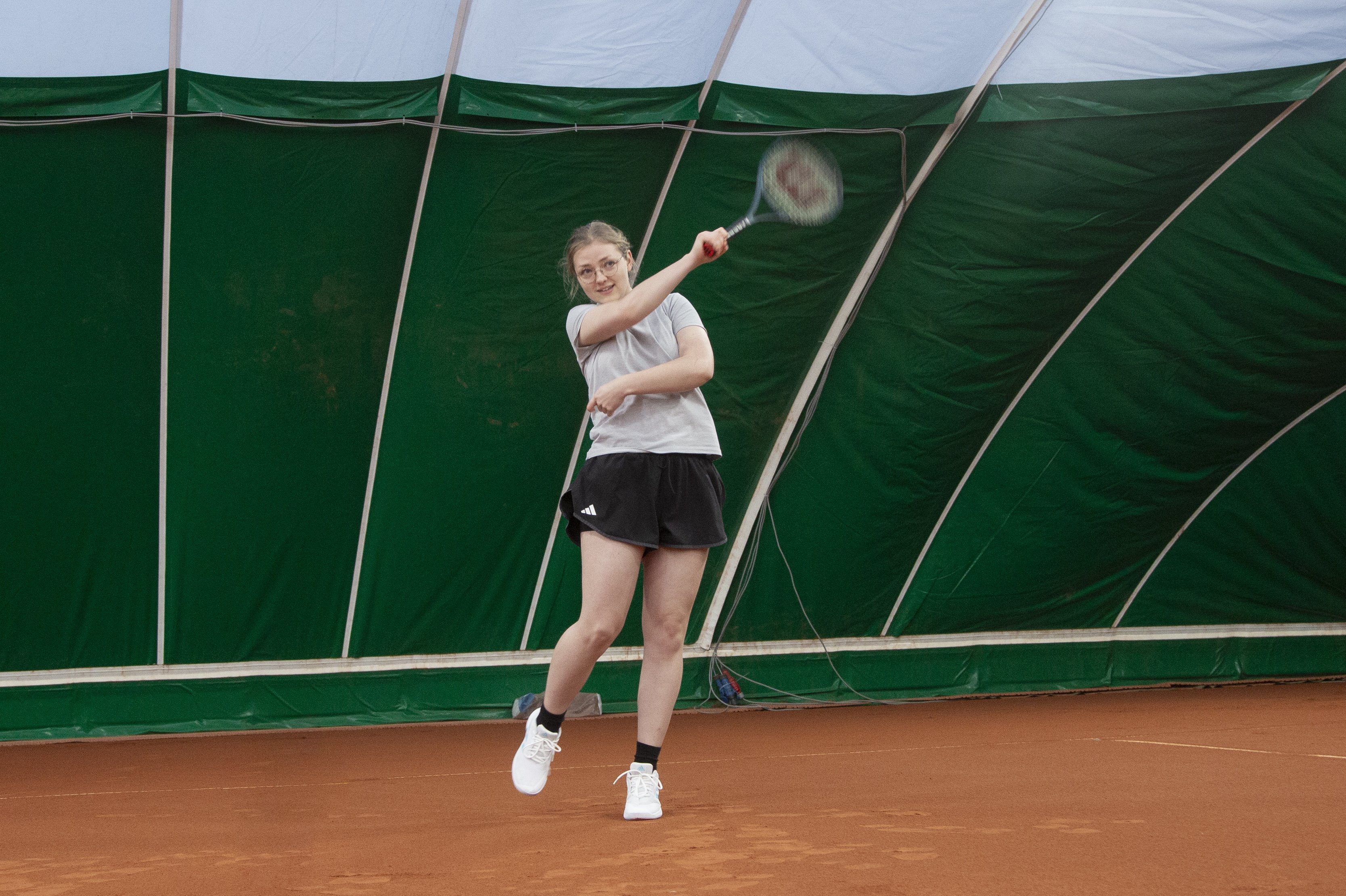 A young woman is playing tennis on a clay court in a covered sports hall. She is wearing sportswear and hits the ball back with her racket.