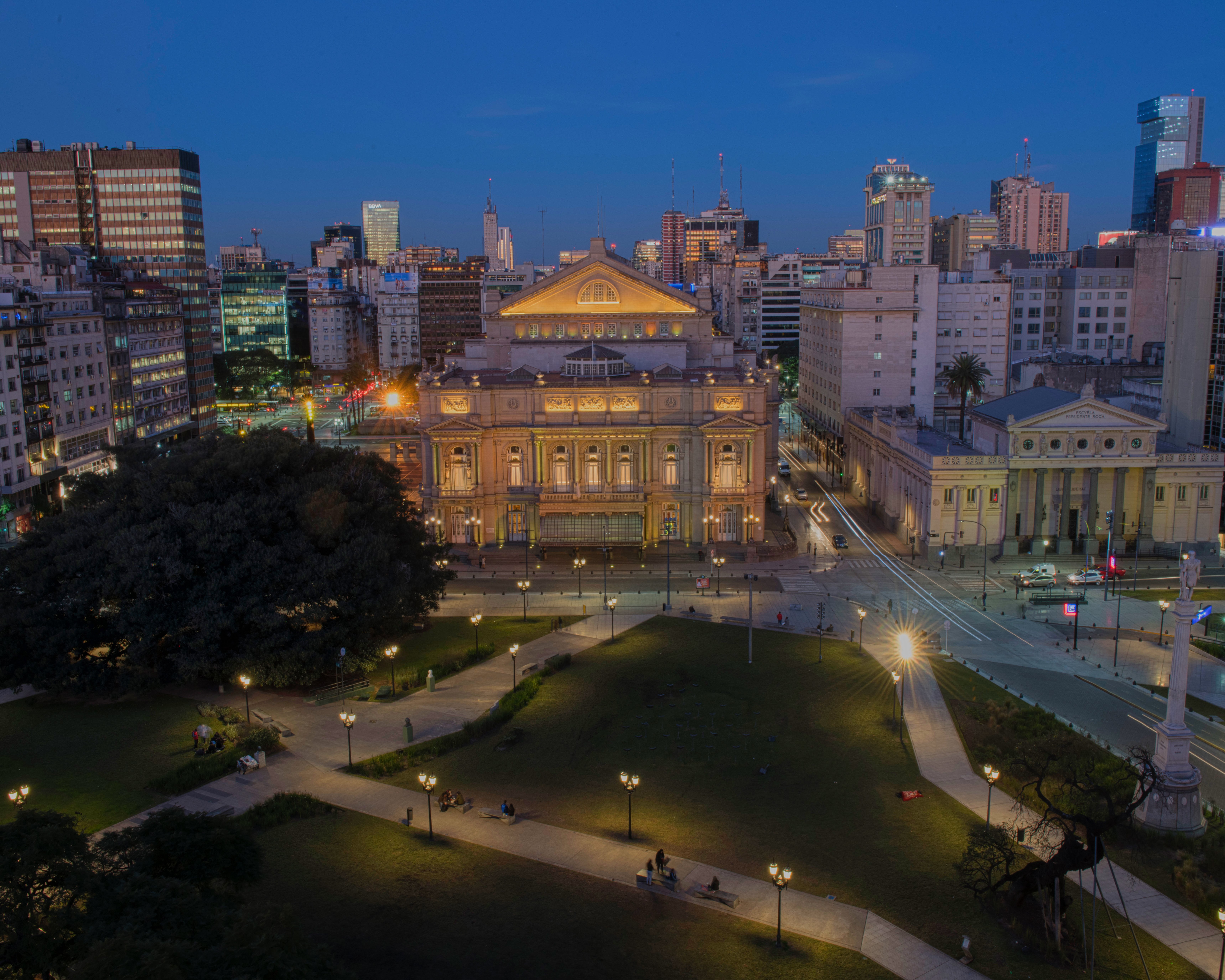 Ein Park in der Abenddämmerung, in dem Menschen auf Bänken sitzen, im Hintergrund sind Stadtgebäude zu sehen, und in der Mitte steht ein großes, beleuchtetes Gebäude im klassischen Stil, umgeben von Straßen und Lichtern.