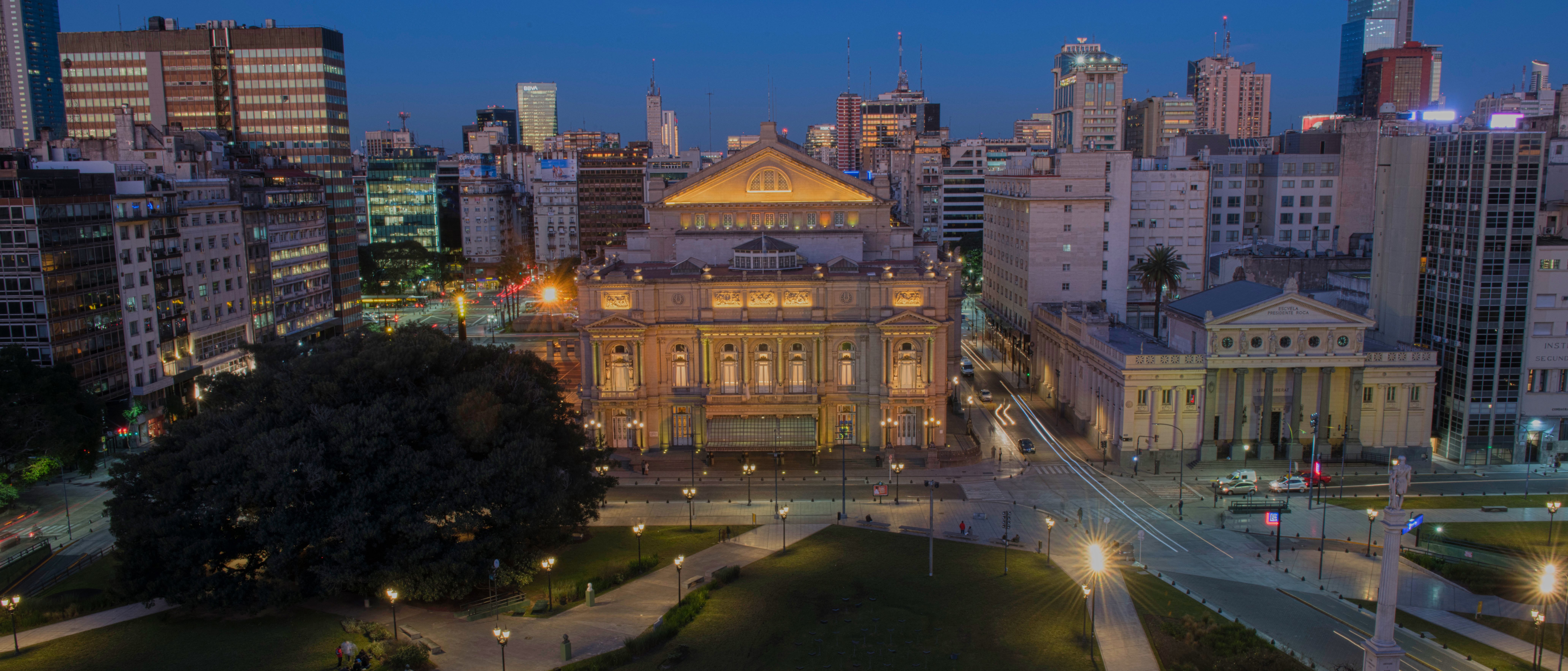 Aerial view of a city park at dusk with a large, illuminated historic building in the center, modern skyscrapers in the background, and scattered people on the grass and pathways.
