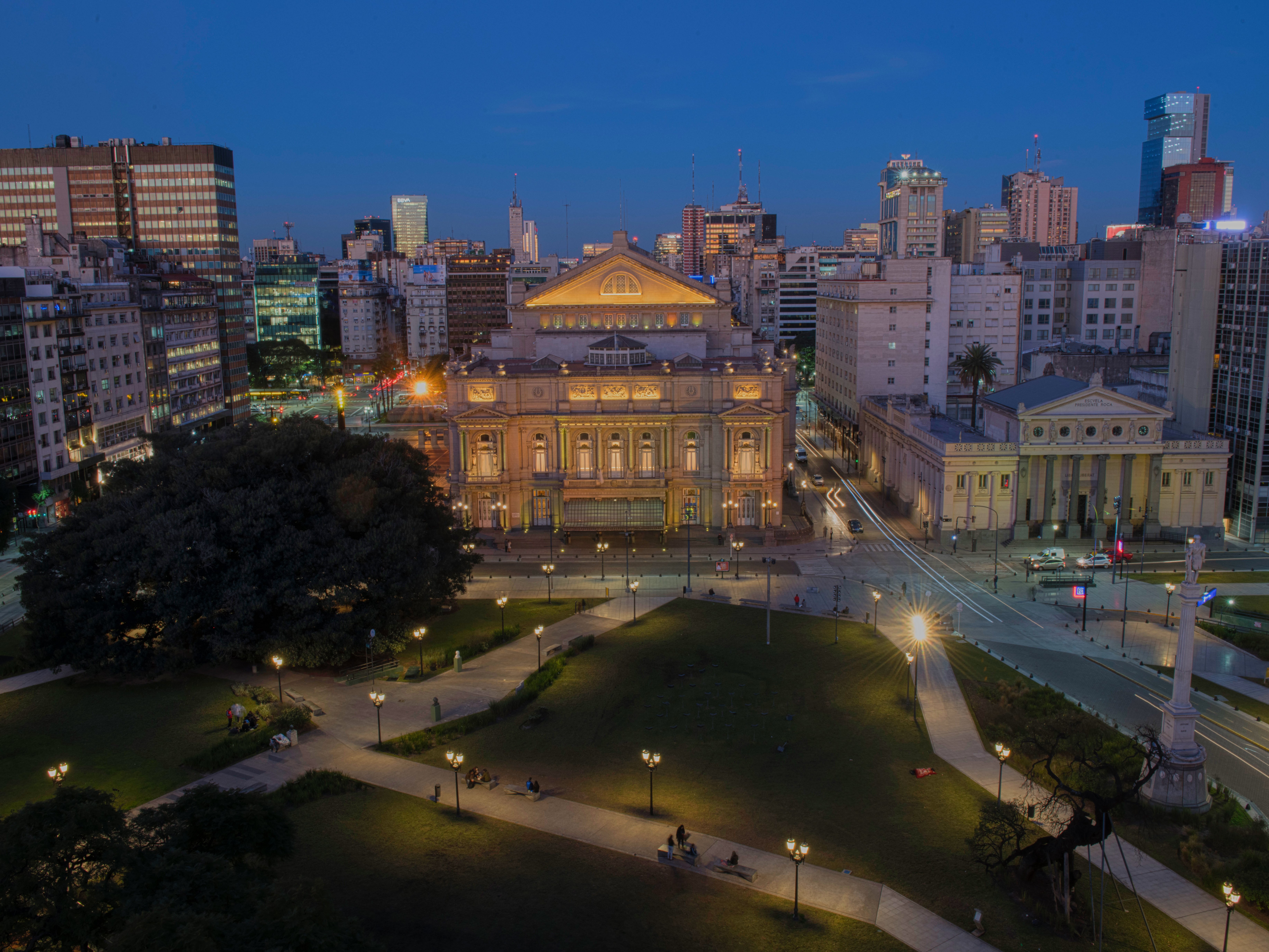 Ein Park in der Abenddämmerung, in dem Menschen auf Bänken sitzen, im Hintergrund sind Stadtgebäude zu sehen, und in der Mitte steht ein großes, beleuchtetes Gebäude im klassischen Stil, umgeben von Straßen und Lichtern.
