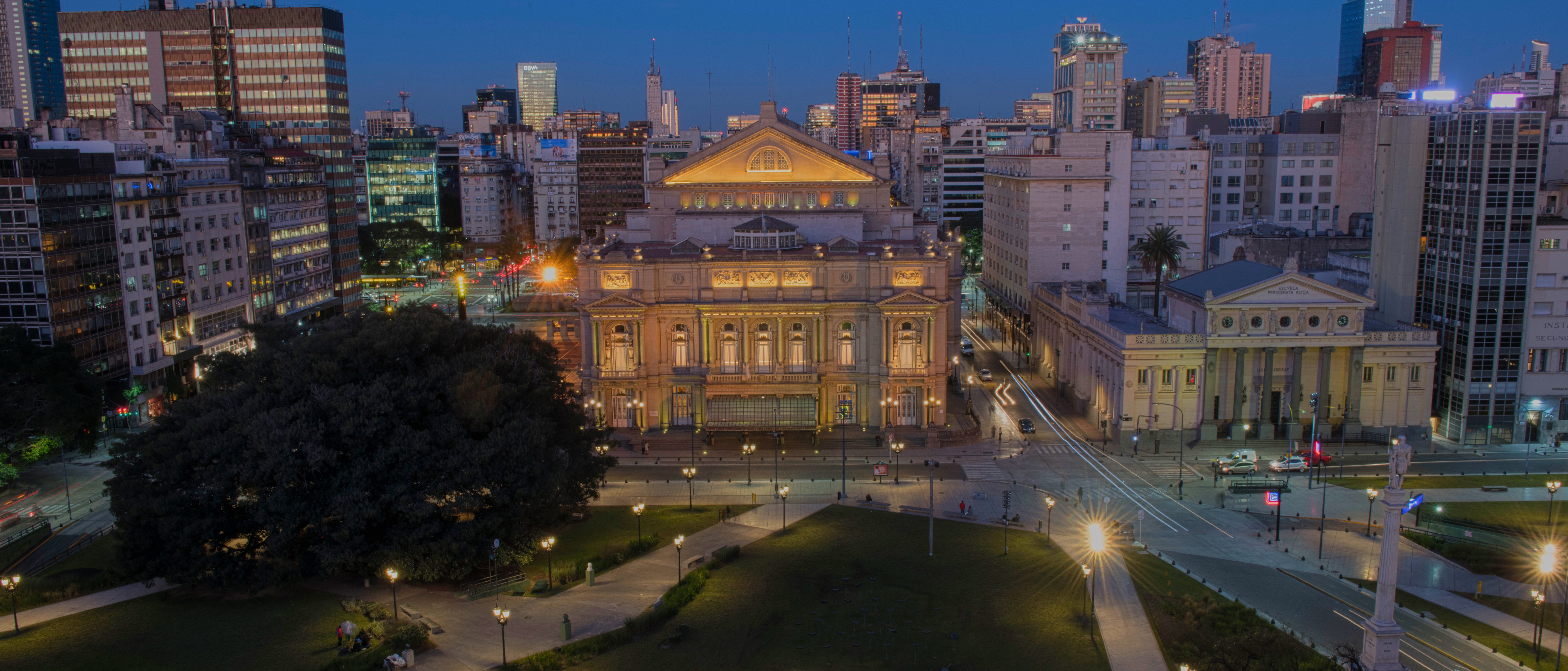 Aerial view of a city park at dusk with a large, illuminated historic building in the center, modern skyscrapers in the background, and scattered people on the grass and pathways.