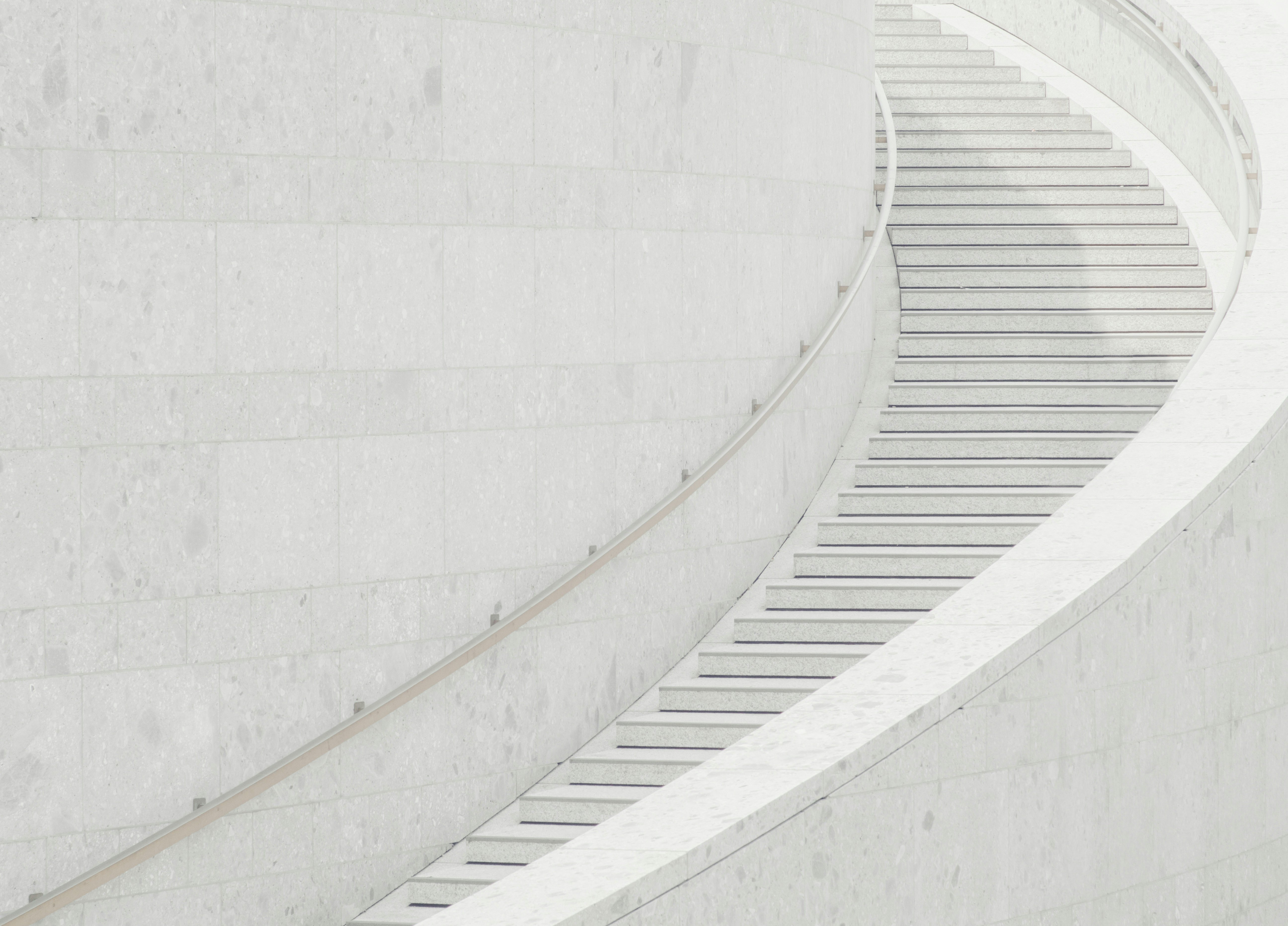 Eine geschwungene Treppe mit weißen Steinstufen und einem glatten Geländer schlängelt sich entlang einer großen, hellen Steinmauer in einem modernen, minimalistischen architektonischen Umfeld.