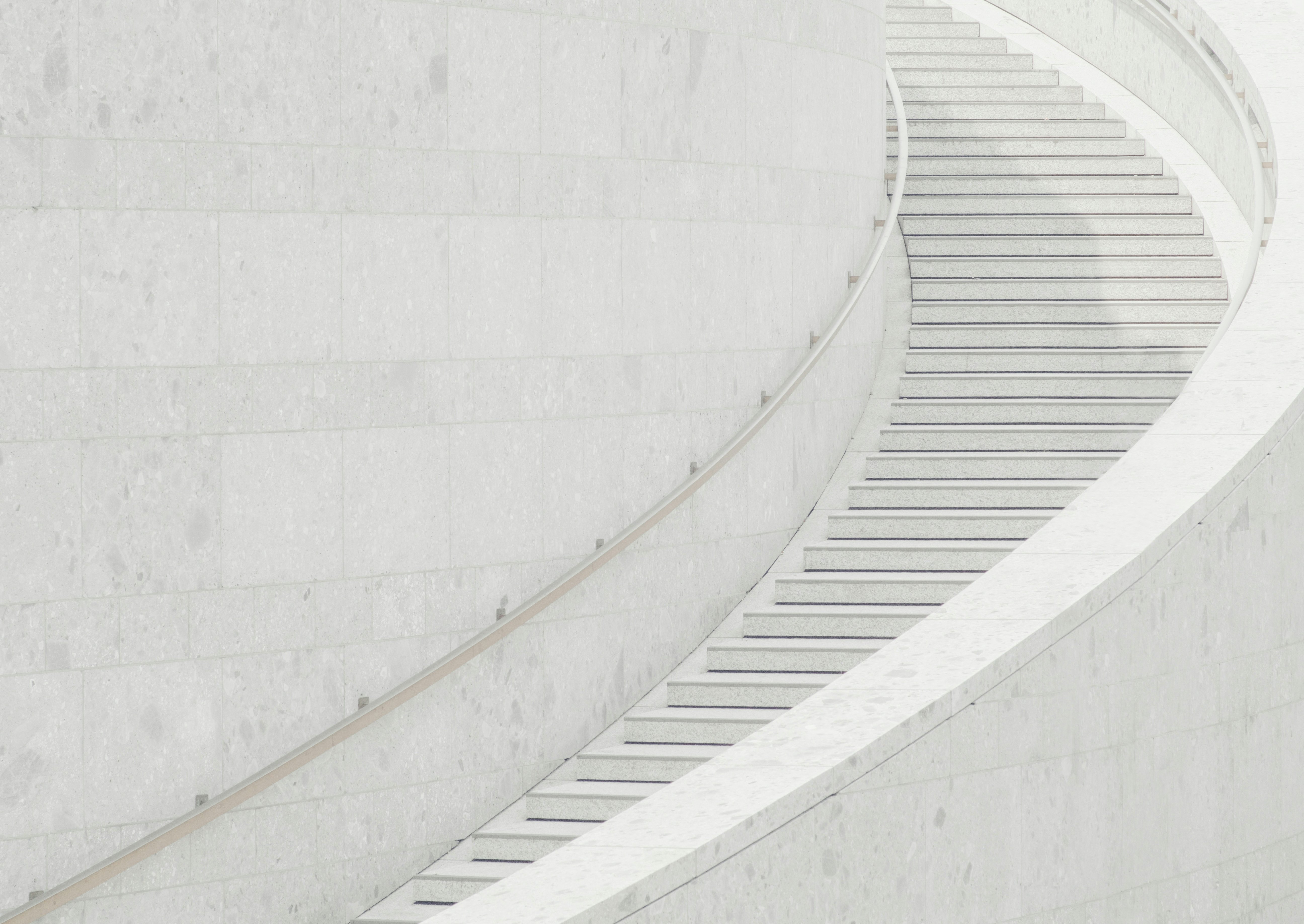A curved, pale stone staircase with a minimalist design winds upward alongside a smooth, light-colored wall. The scene is bright and modern, with clean lines and soft shadows.