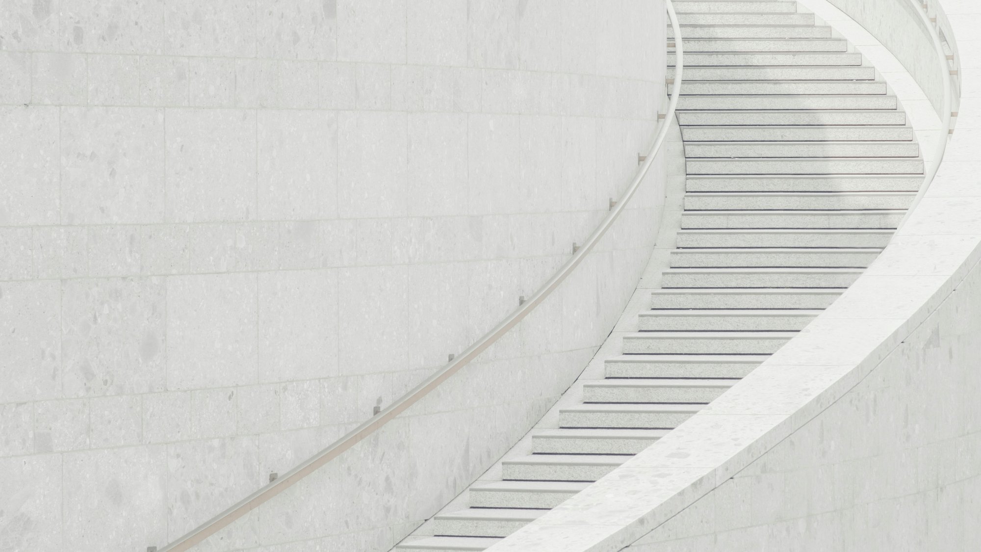 A curved, pale stone staircase with a minimalist design winds upward alongside a smooth, light-colored wall. The scene is bright and modern, with clean lines and soft shadows.