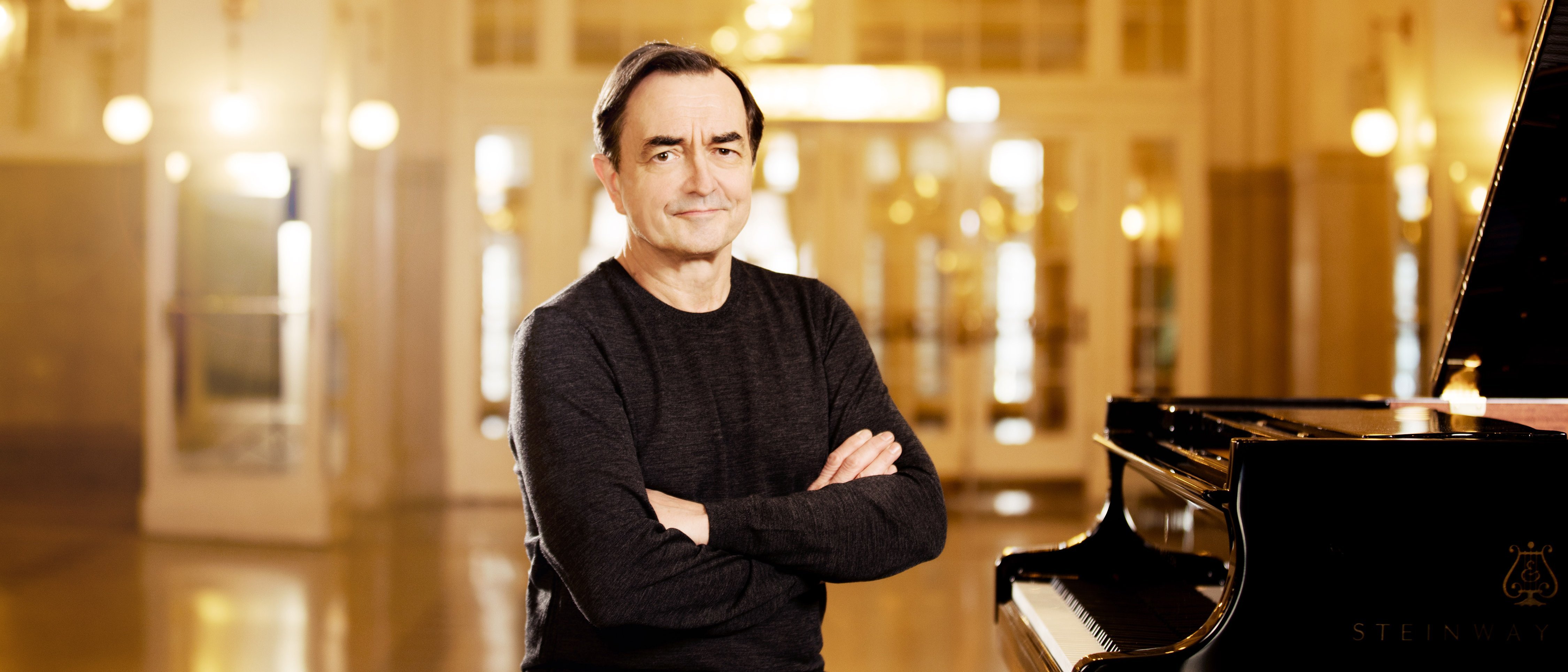Pierre-Laurant Aimard sits in front of a grand piano in a large, illuminated room