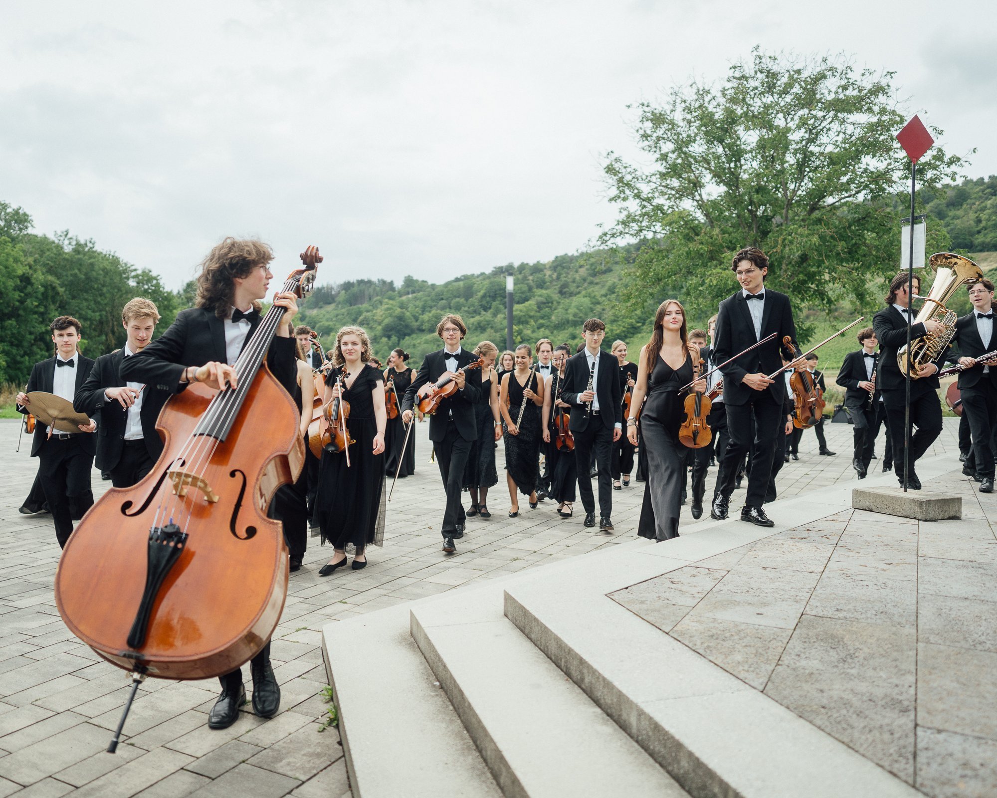 A large group of young musicians in formal black attire walk with their instruments, including a double bass, violins and brass instruments, on a paved area surrounded by greenery.