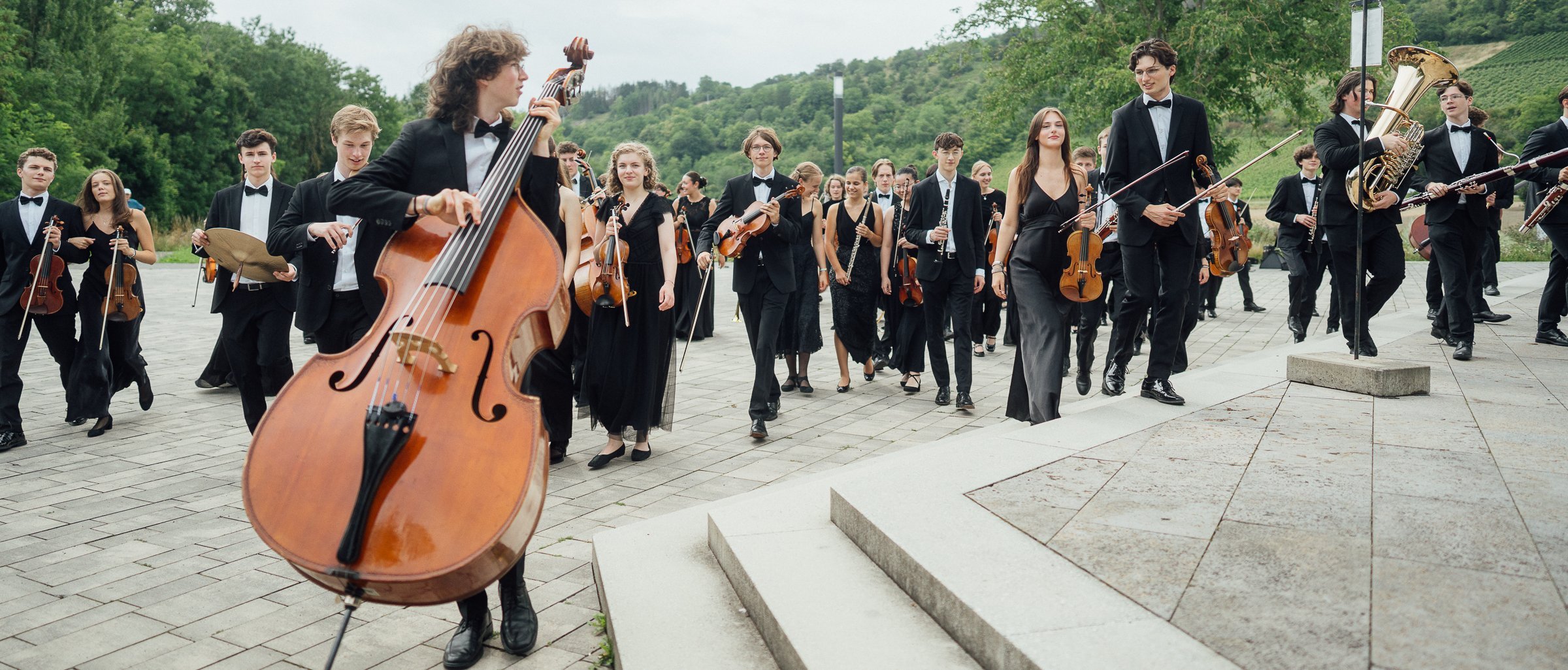 Eine große Gruppe junger Musiker in formeller schwarzer Kleidung geht mit ihren Instrumenten, darunter ein Kontrabass, Geigen und Blechbläser, auf einer gepflasterten Fläche im Grünen.