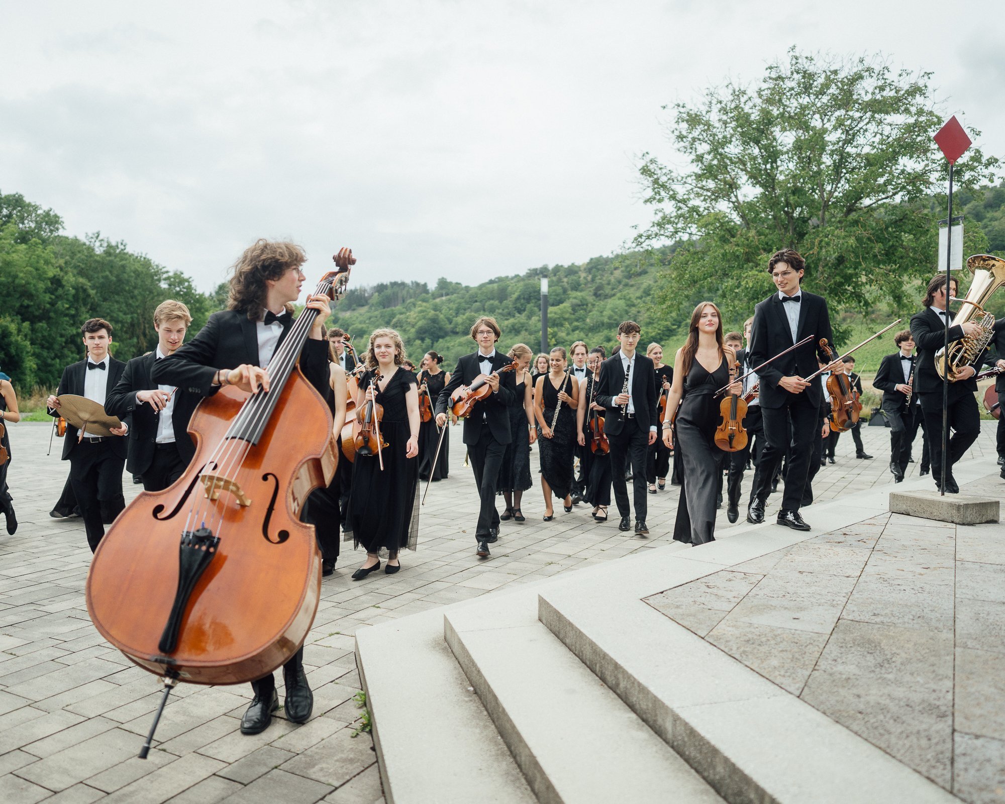 Eine große Gruppe junger Musiker in formeller schwarzer Kleidung geht mit ihren Instrumenten, darunter ein Kontrabass, Geigen und Blechbläser, auf einer gepflasterten Fläche im Grünen.