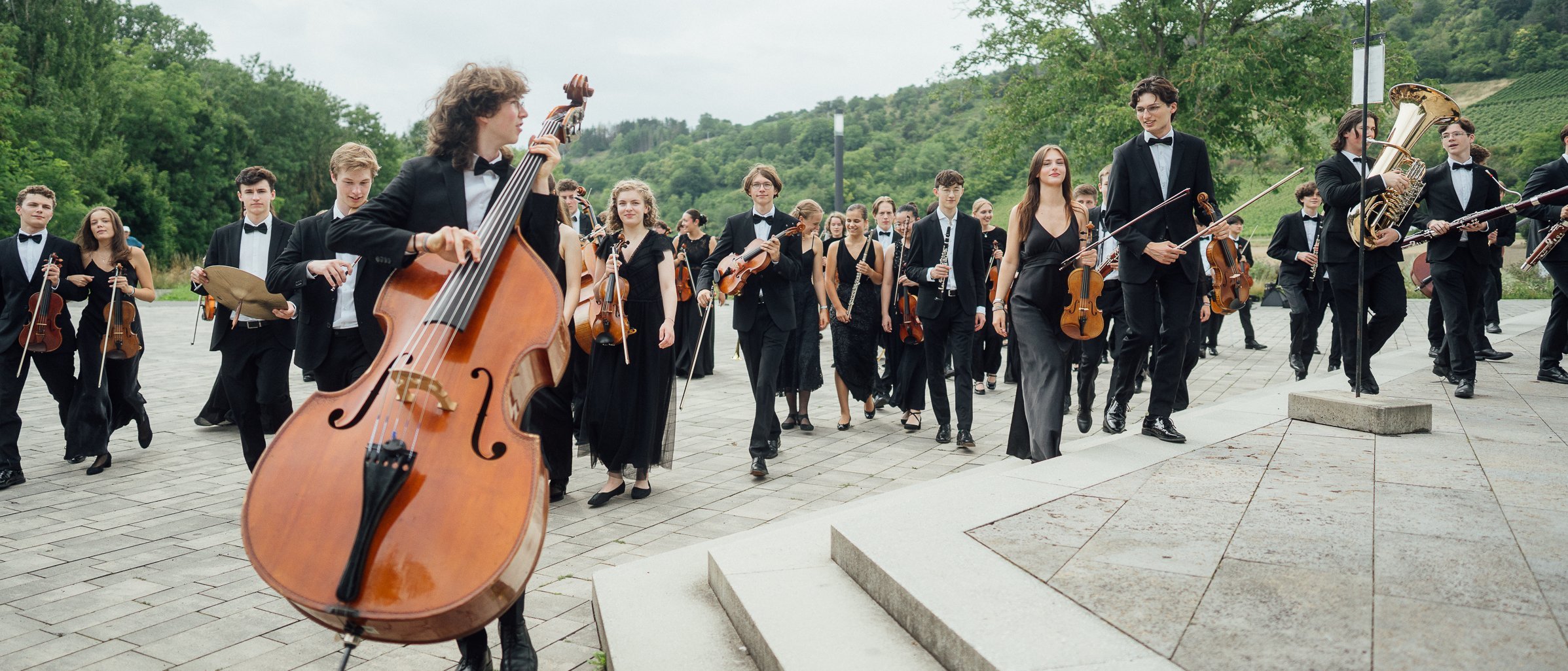 Eine große Gruppe junger Musiker in formeller schwarzer Kleidung geht mit ihren Instrumenten, darunter ein Kontrabass, Geigen und Blechbläser, auf einer gepflasterten Fläche im Grünen.