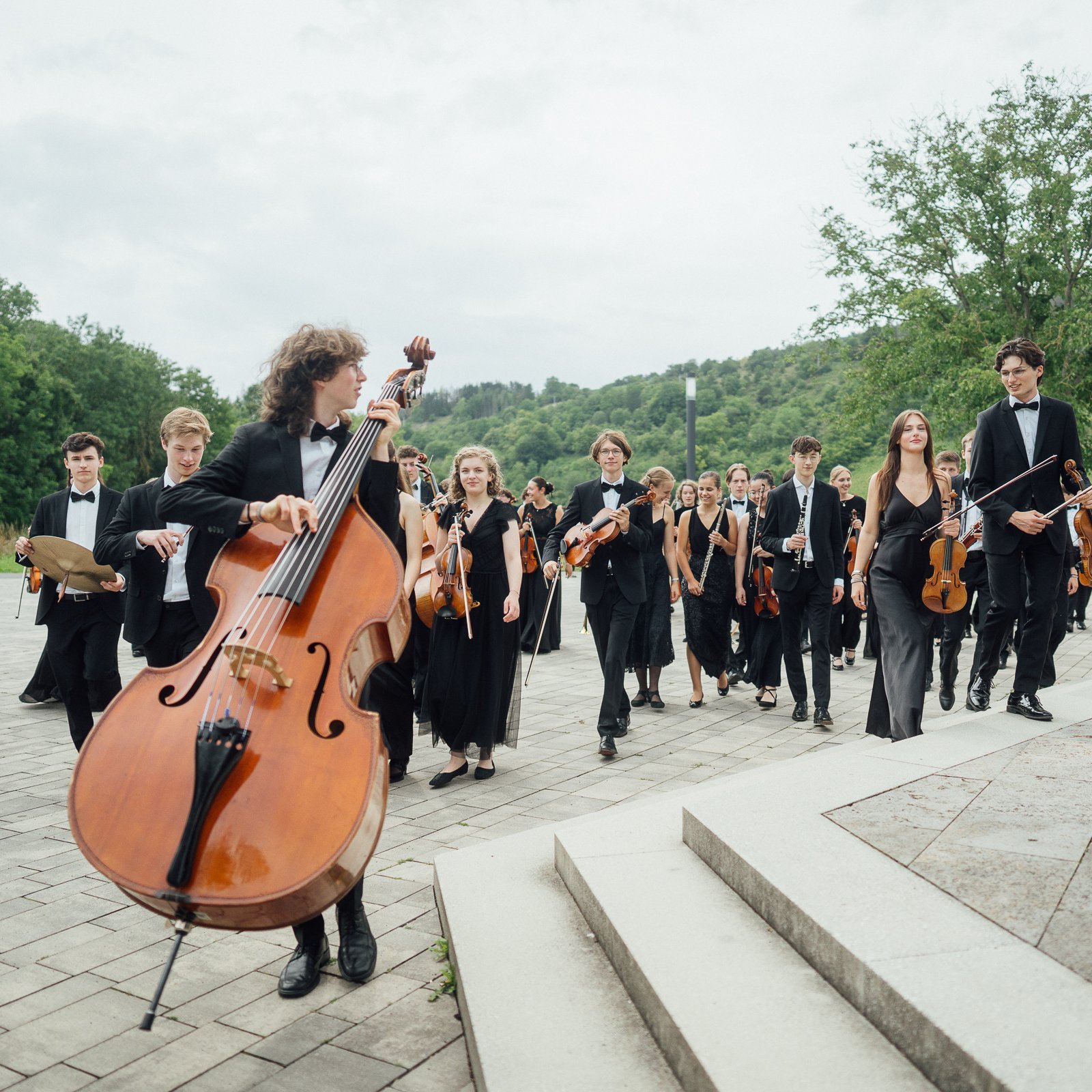 Eine große Gruppe junger Musiker in formeller schwarzer Kleidung geht mit ihren Instrumenten, darunter ein Kontrabass, Geigen und Blechbläser, auf einer gepflasterten Fläche im Grünen.