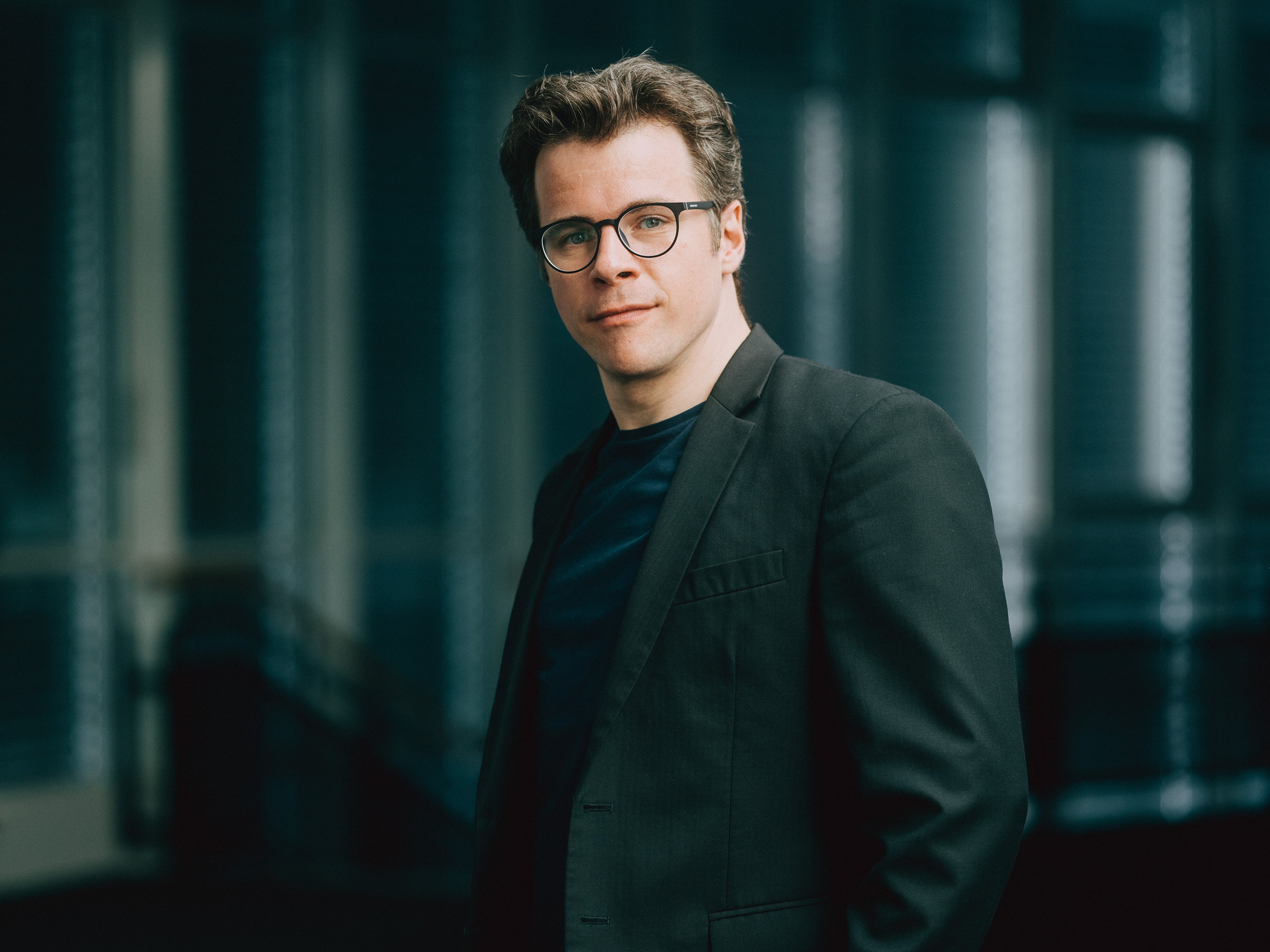 Jakub Hrůša mit Brille und dunklem Anzug blickt mit einem leichten Lächeln in die Kamera. Er steht in einer Innenumgebung mit sanftem Licht und unscharfem Hintergrund.