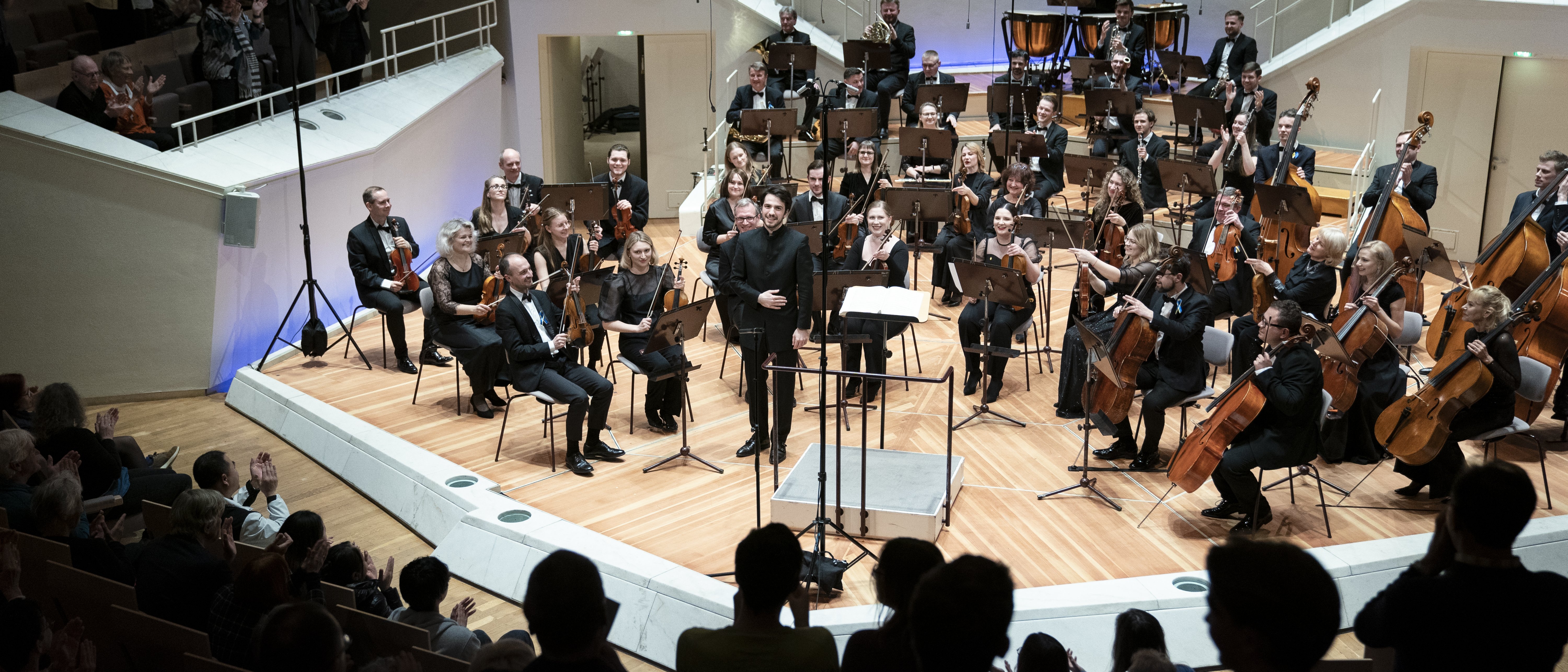 The Kyiv Symphony Orchestra sits on stage during the final applause. Conductor Christian Blex takes a bow. Clapping people can be seen in the foreground.