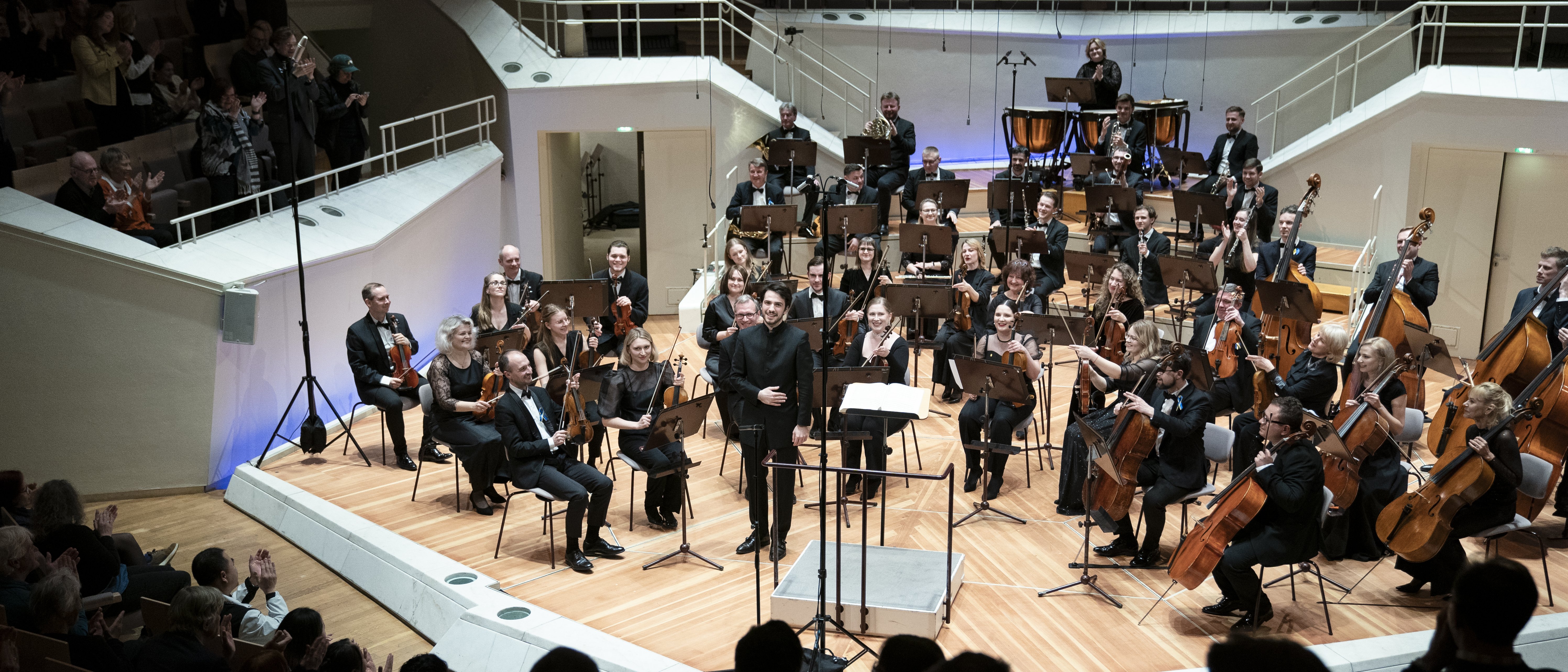 Das Kyiv Symphony Orchestra sitzt auf der Bühne beim Schlussapplaus. Der Dirigent Christian Blex verbeugt sich. Im Vordergrund sind klatschende Menschen zu sehen.