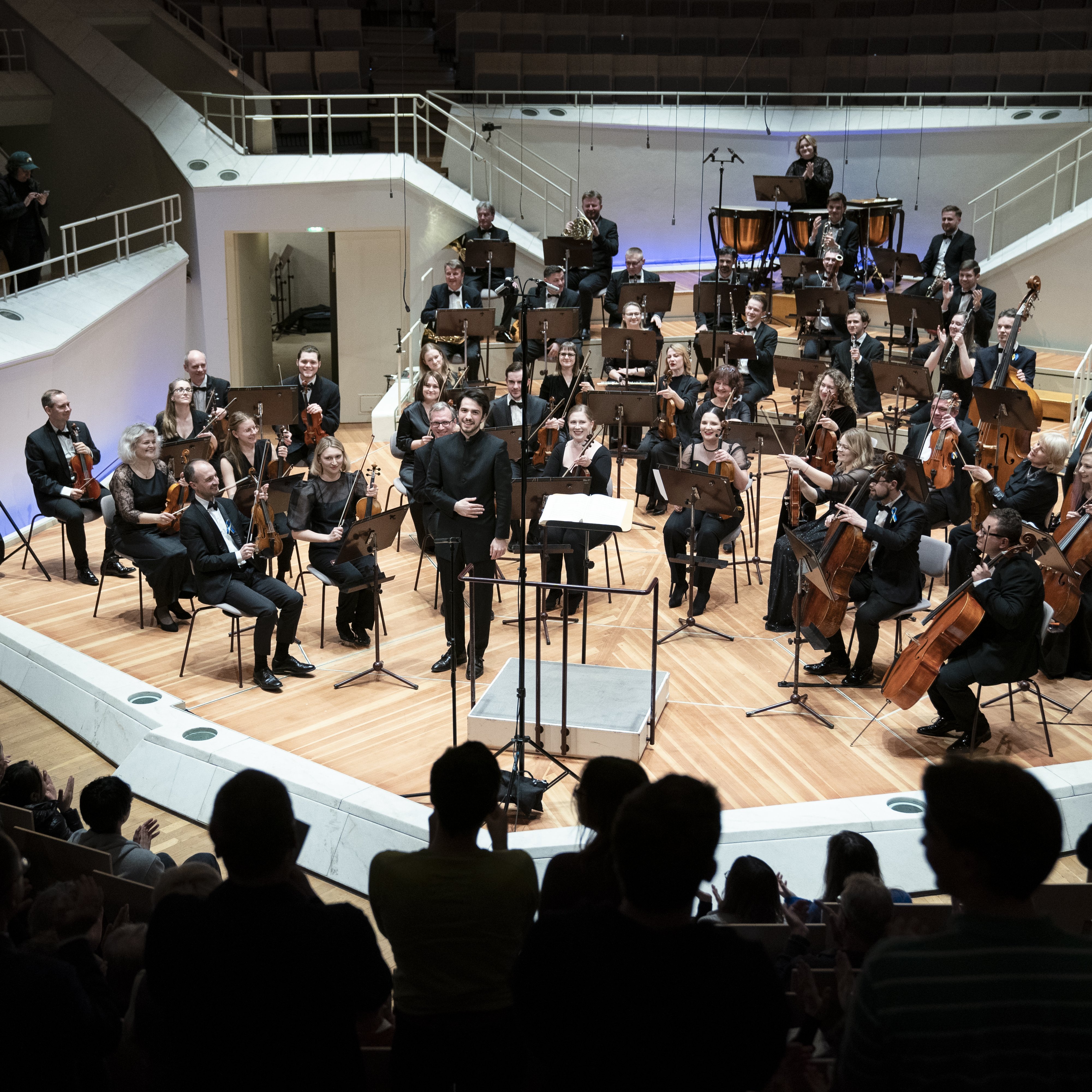 Das Kyiv Symphony Orchestra sitzt auf der Bühne beim Schlussapplaus. Der Dirigent Christian Blex verbeugt sich. Im Vordergrund sind klatschende Menschen zu sehen.