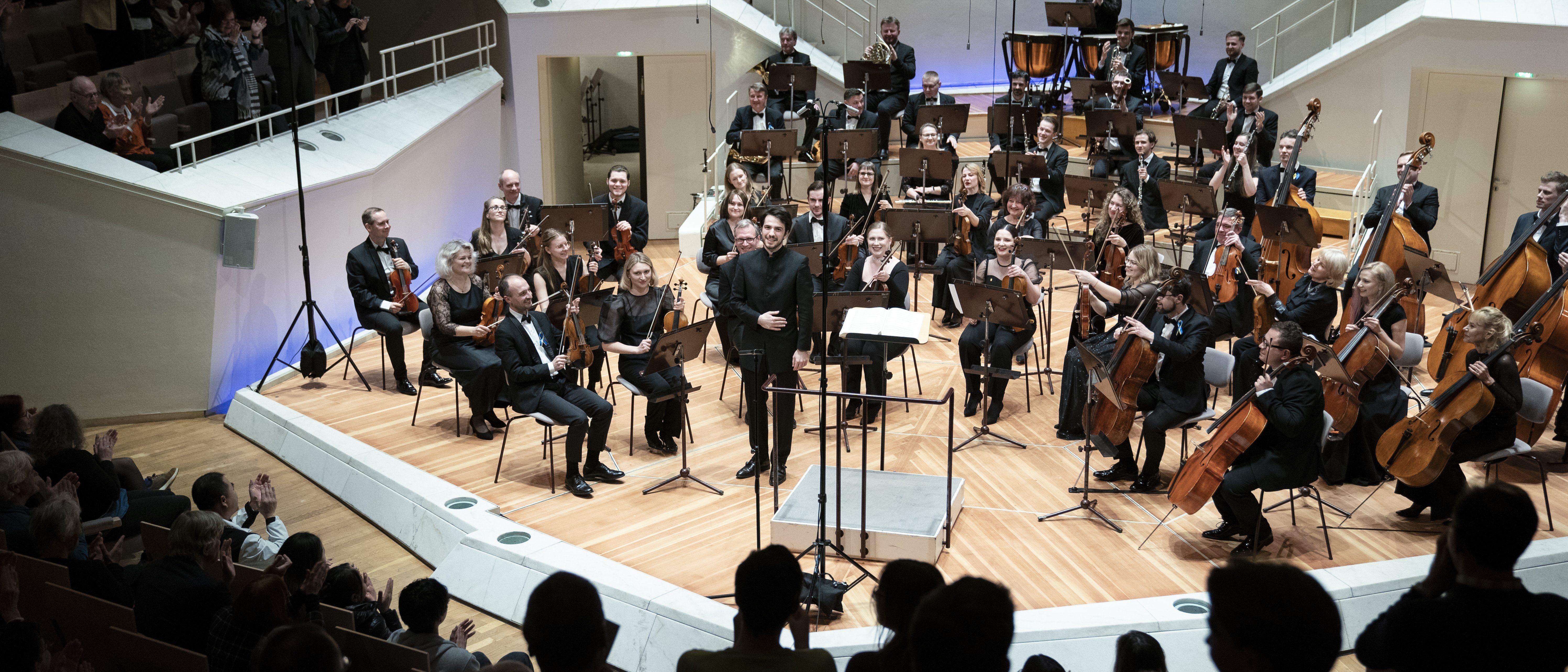 Das Kyiv Symphony Orchestra sitzt auf der Bühne beim Schlussapplaus. Der Dirigent Christian Blex verbeugt sich. Im Vordergrund sind klatschende Menschen zu sehen.