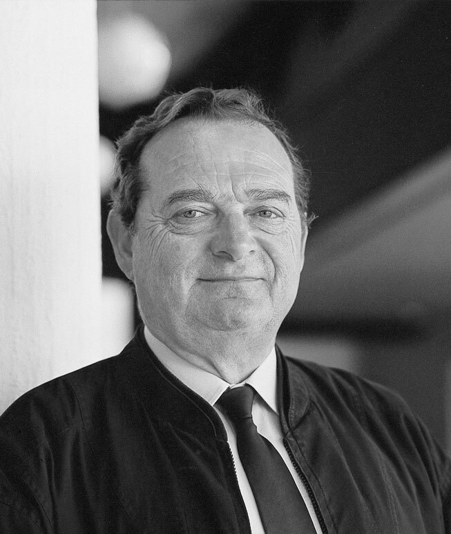Black and white portrait of an older man in a suit and tie, standing indoors, smiling slightly at the camera with short hair; the background is softly blurred.