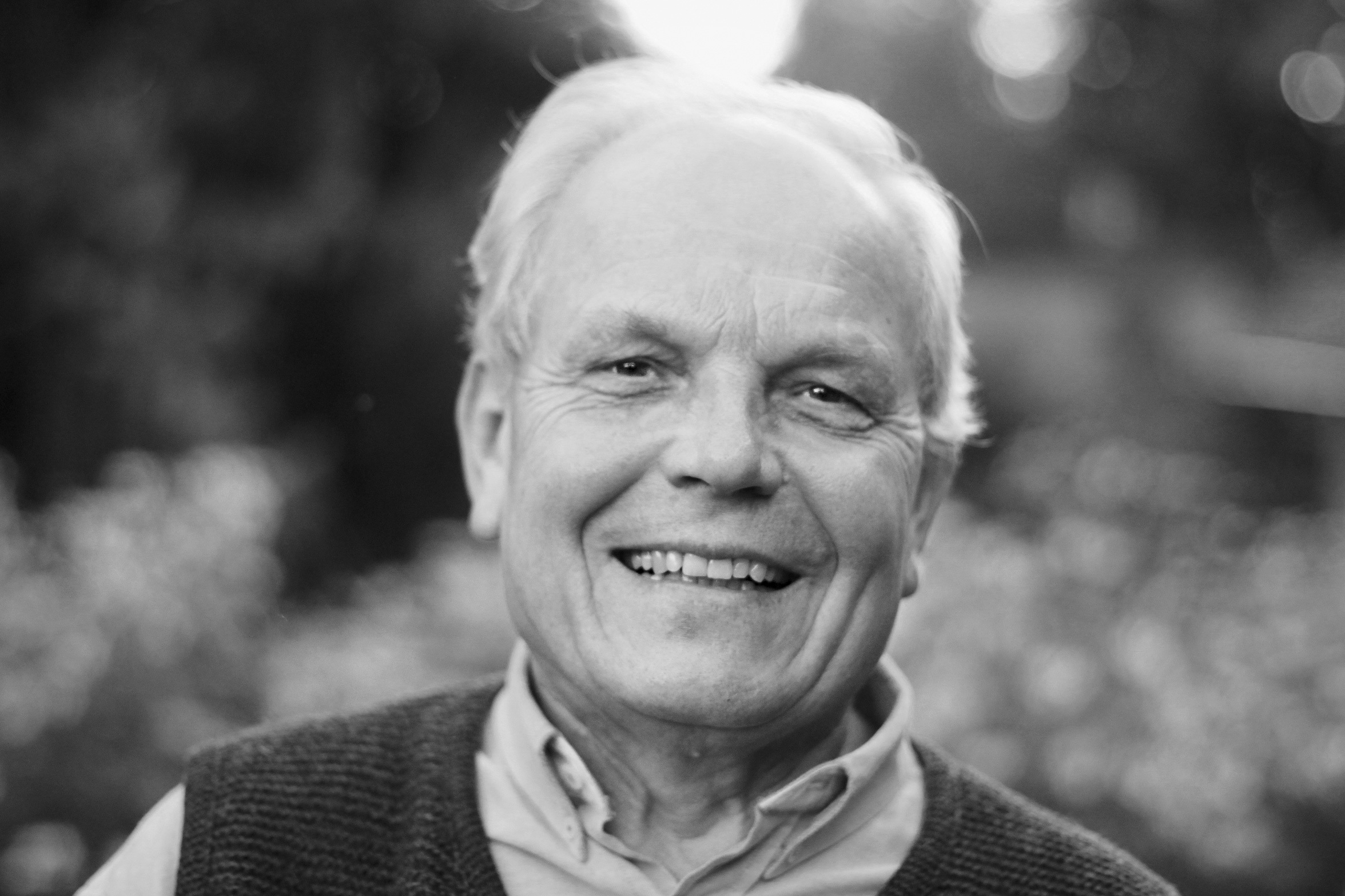 An elderly man with light hair smiles warmly at the camera. He is wearing a shirt with a knitted waistcoat over it. The background is blurred and shows a natural setting with trees and light reflections.