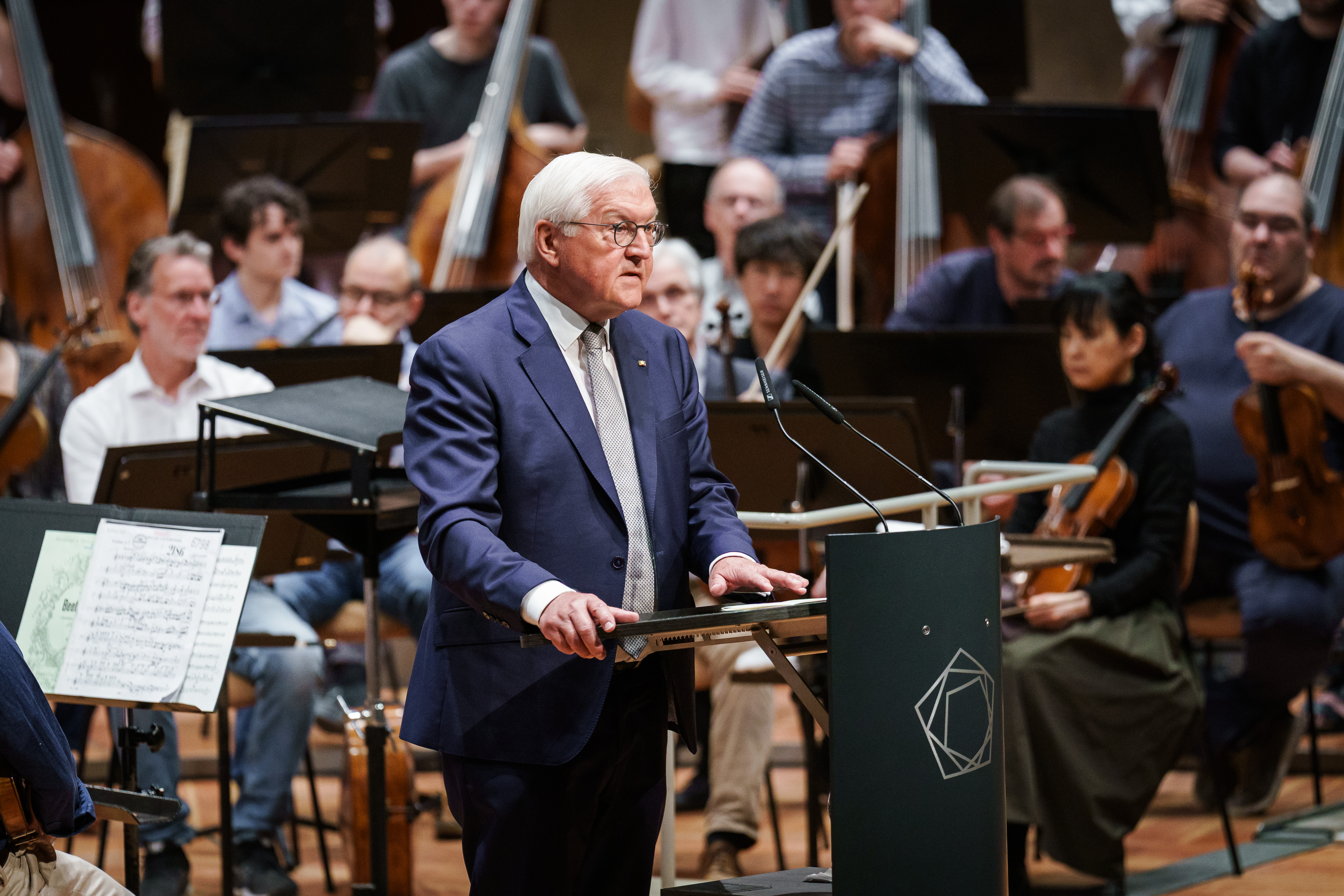 Frank Walter Steinmeier in einem blauen Anzug spricht auf einem Podium vor einem Orchester, hinter ihm sitzen Musiker, die ihre Instrumente halten, und im Vordergrund sind Noten zu sehen.