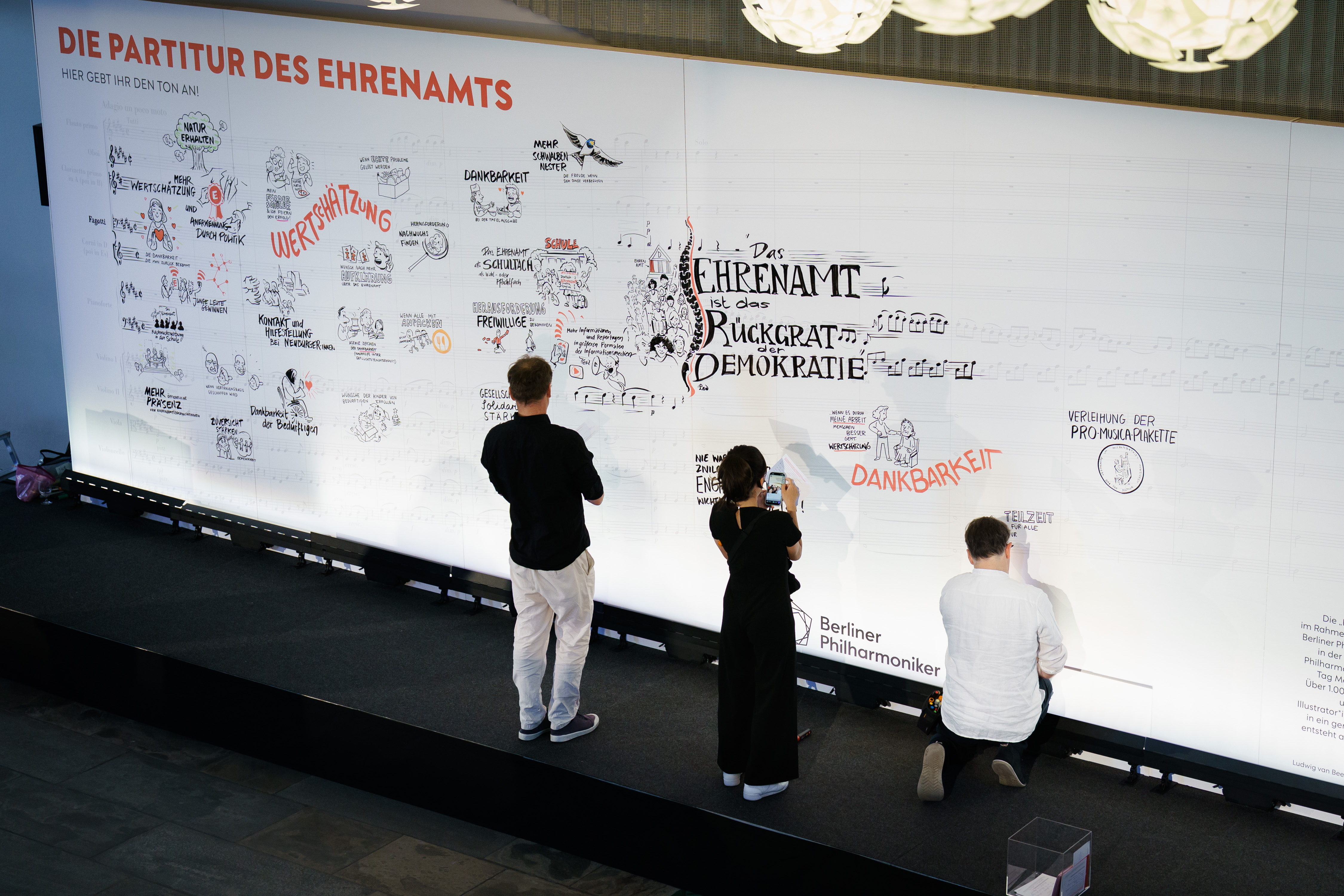 Drei Personen illustrieren und schreiben auf einem großen Whiteboard mit deutschen Texten und Zeichnungen bei einer Veranstaltung mit dem Titel Die Partitur des Ehrenamts in einem gut beleuchteten Innenraum.