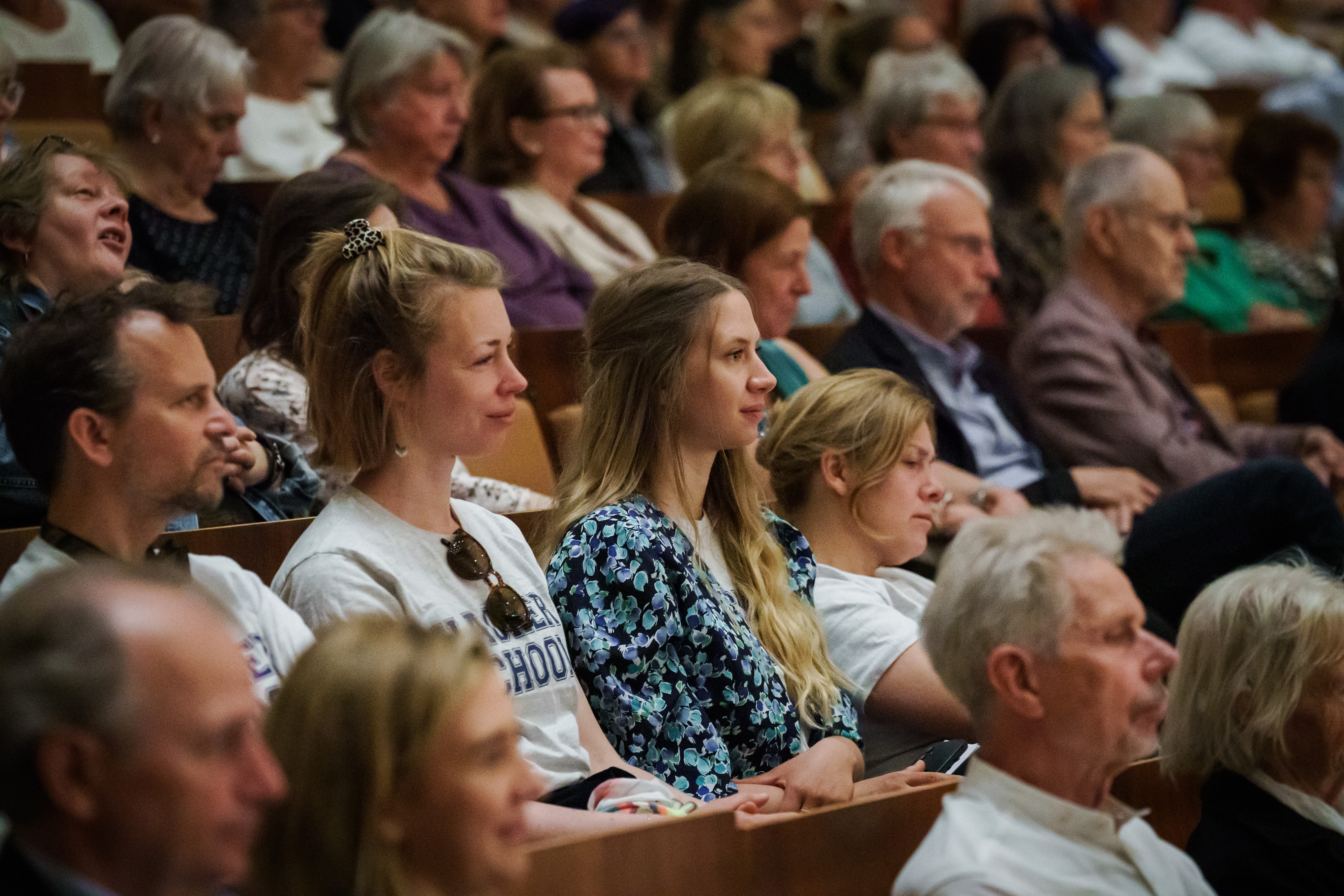 Ein bunt gemischtes Publikum aus Männern und Frauen, Jung und Alt, sitzt aufmerksam in den Reihen eines Hörsaals und konzentriert sich auf ein nicht sichtbares Ereignis oder einen Redner im Vordergrund.