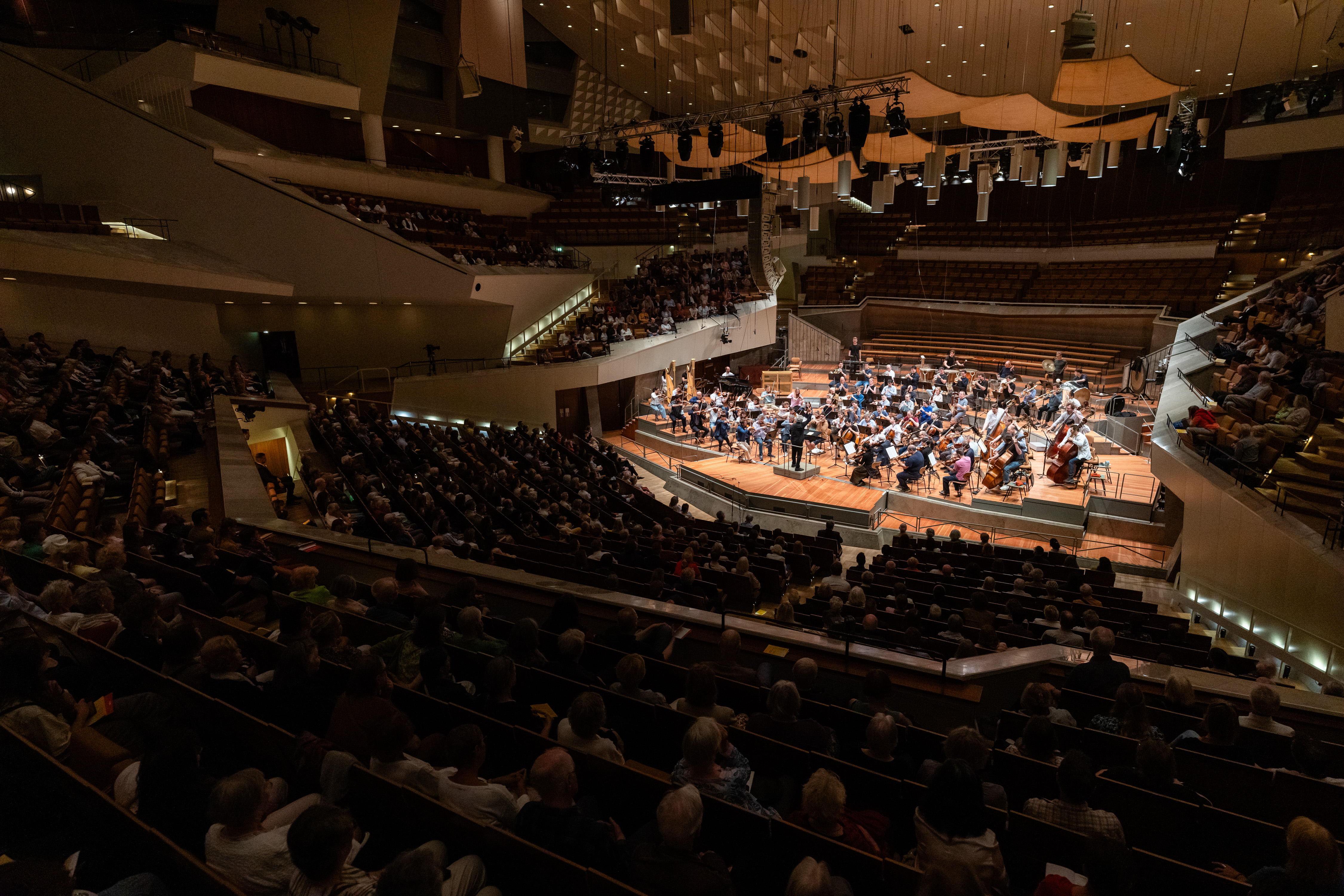 Blick auf einen großen Konzertsaal, der mit Zuschauern gefüllt ist, die einem Orchester auf einer gut beleuchteten Bühne zusehen, mit abgestuften Sitzreihen und modernen architektonischen Details, die überall im Saal zu sehen sind.