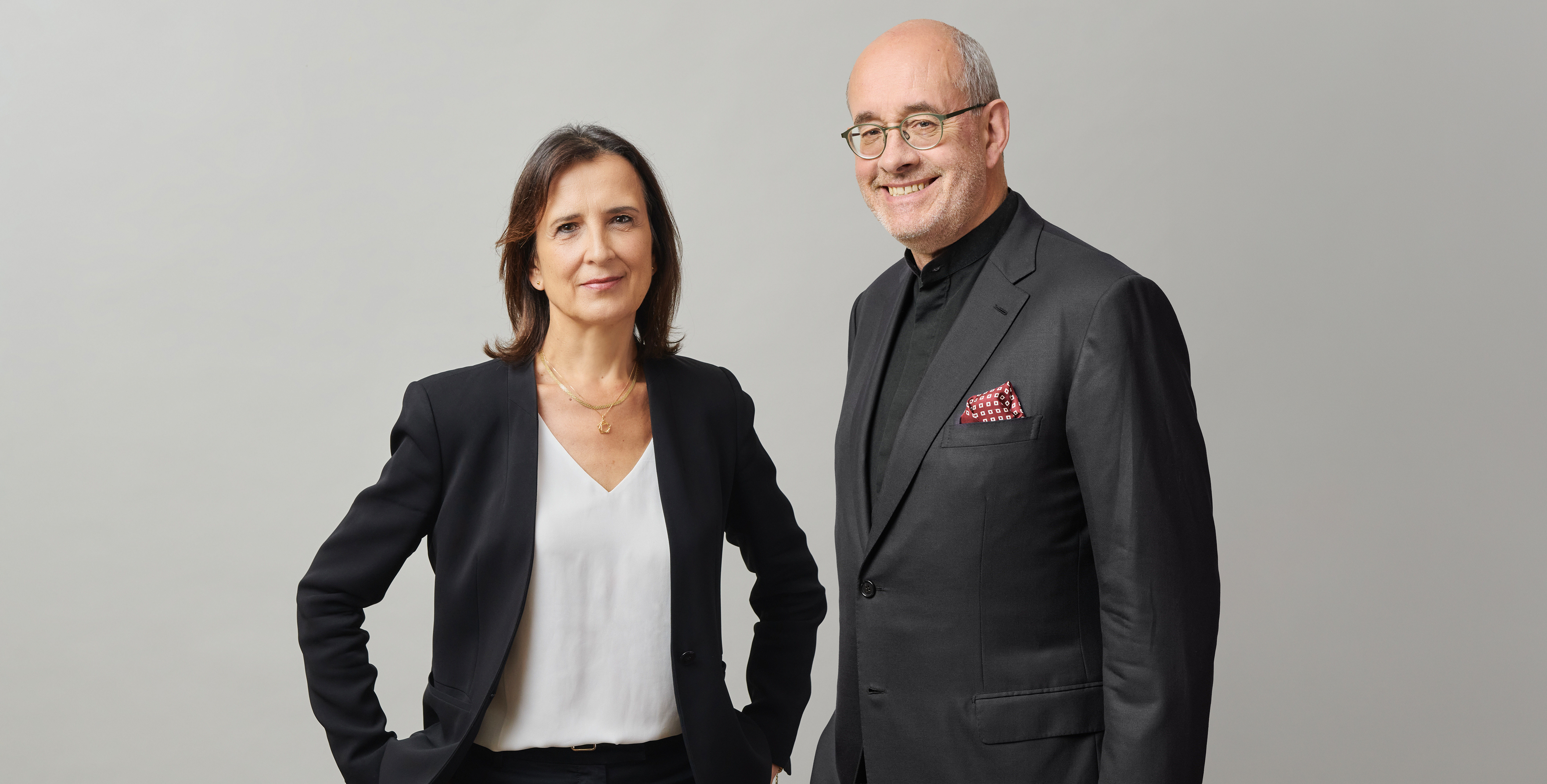 A woman and a man in business attire stand side by side against a plain light gray background, both smiling at the camera.