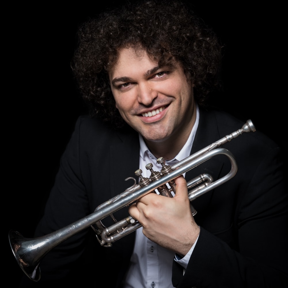 A smiling man with curly hair, wearing a dark suit and white shirt, holds a silver trumpet against a black background.