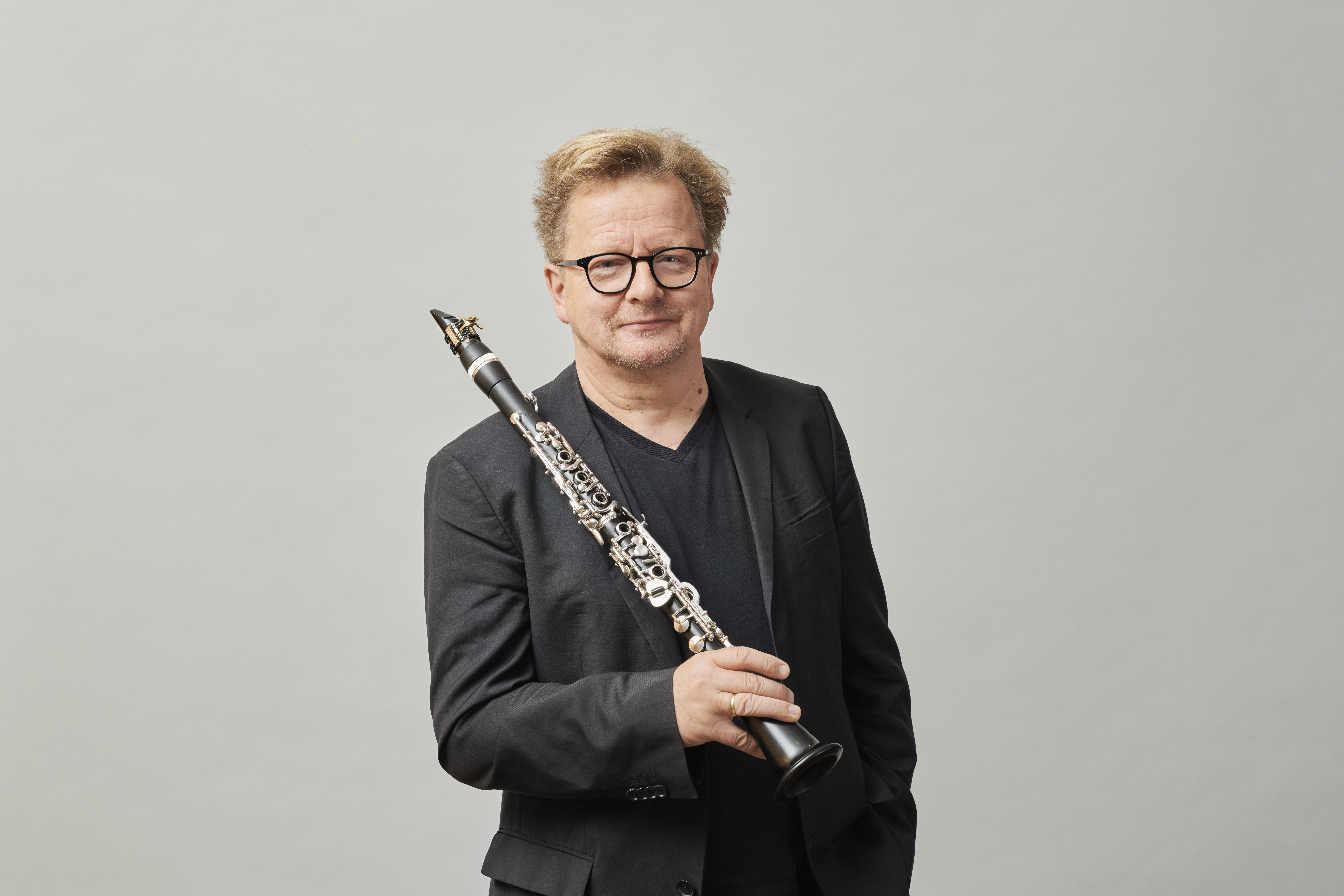 Wenzel Fuchs with short, light-coloured hair and glasses, wearing a black blazer and a black shirt, stands in front of a plain background and holds a clarinet upright in his right hand.