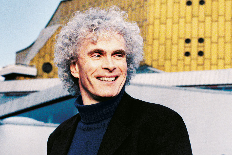 Ein lächelnder Simon Rattle mit lockigem grauem Haar, der einen dunklen Rollkragenpullover und eine Jacke trägt, steht im Freien vor einem modernen gelb-weißen Gebäude.