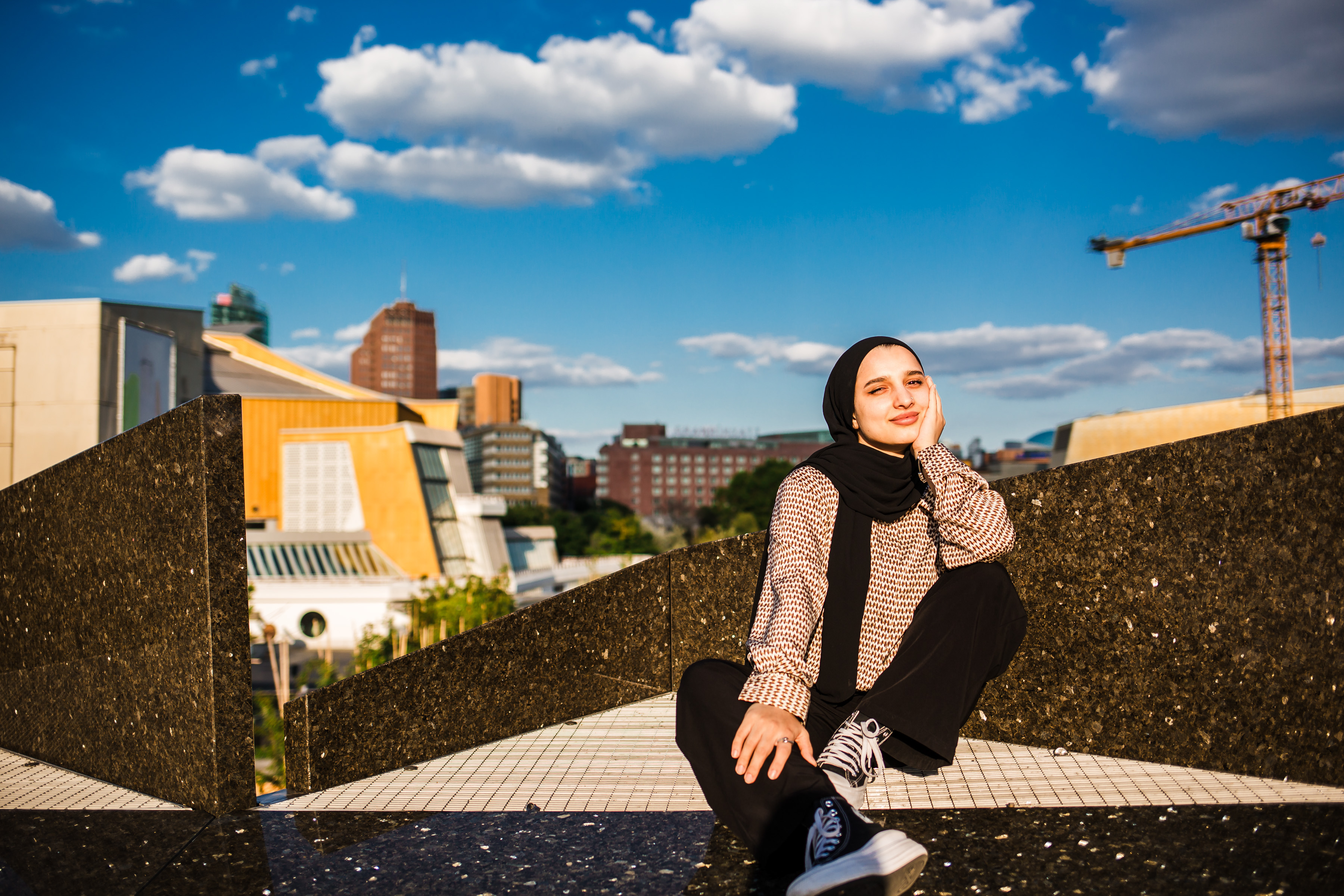 Eine Frau in Hidschab und Freizeitkleidung sitzt auf einer geometrischen Steinstruktur und legt lächelnd die Hand auf ihr Kinn. Im Hintergrund sind städtische Gebäude, ein Kran und ein blauer Himmel mit Wolken zu sehen.
