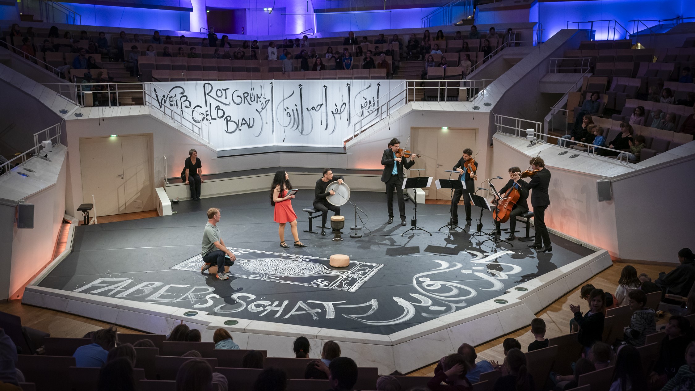 On stage, musicians are playing instruments, and a woman in a red dress holds cue cards. The stage floor is black, with some drawings made in chalk on it.