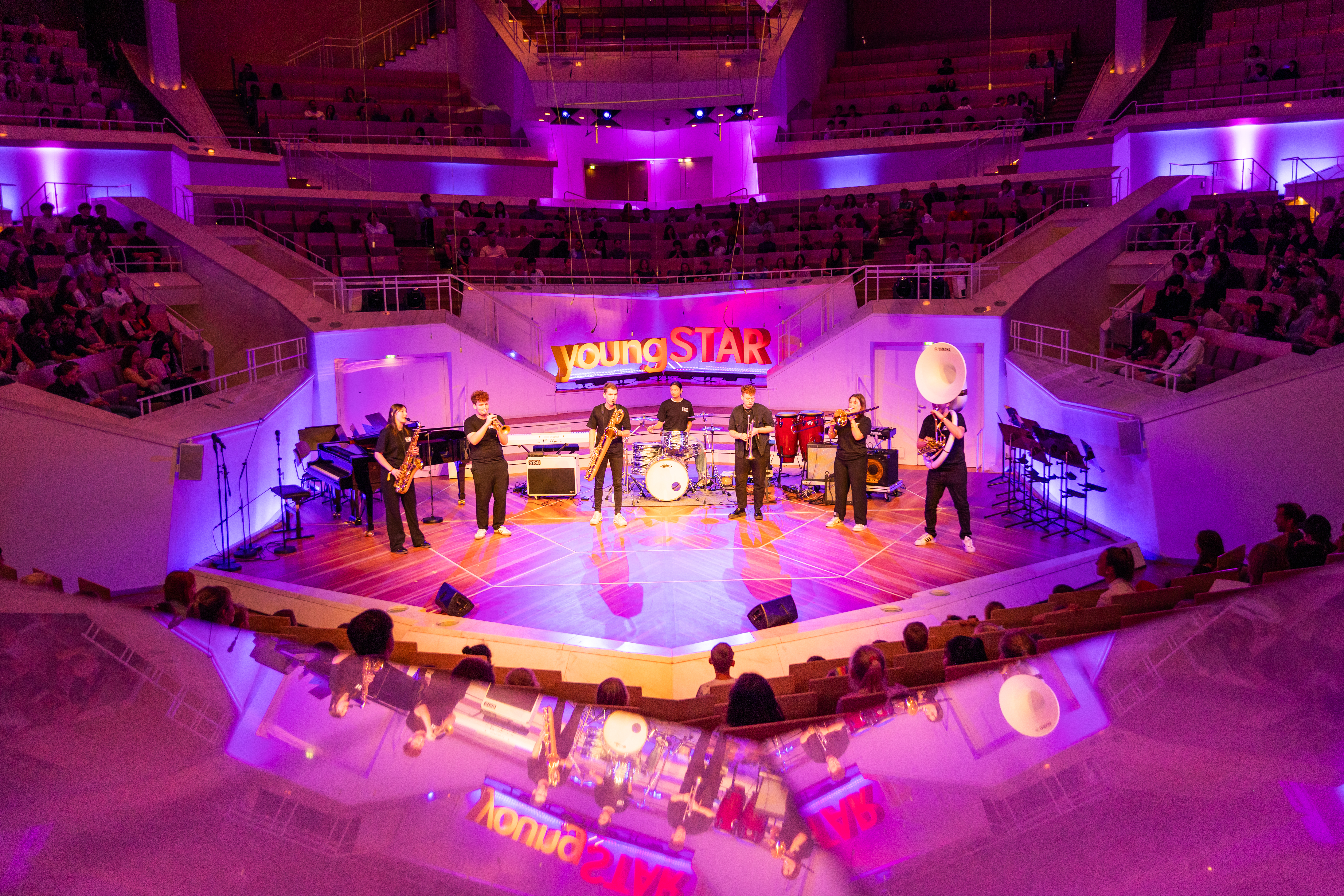 In einem Konzertsaal tritt eine Band auf einer hell erleuchteten Bühne mit violetter Beleuchtung auf. Das Publikum sitzt in gestuften Reihen, und hinter den Musikern ist ein „youngSTAR»-Banner zu sehen.