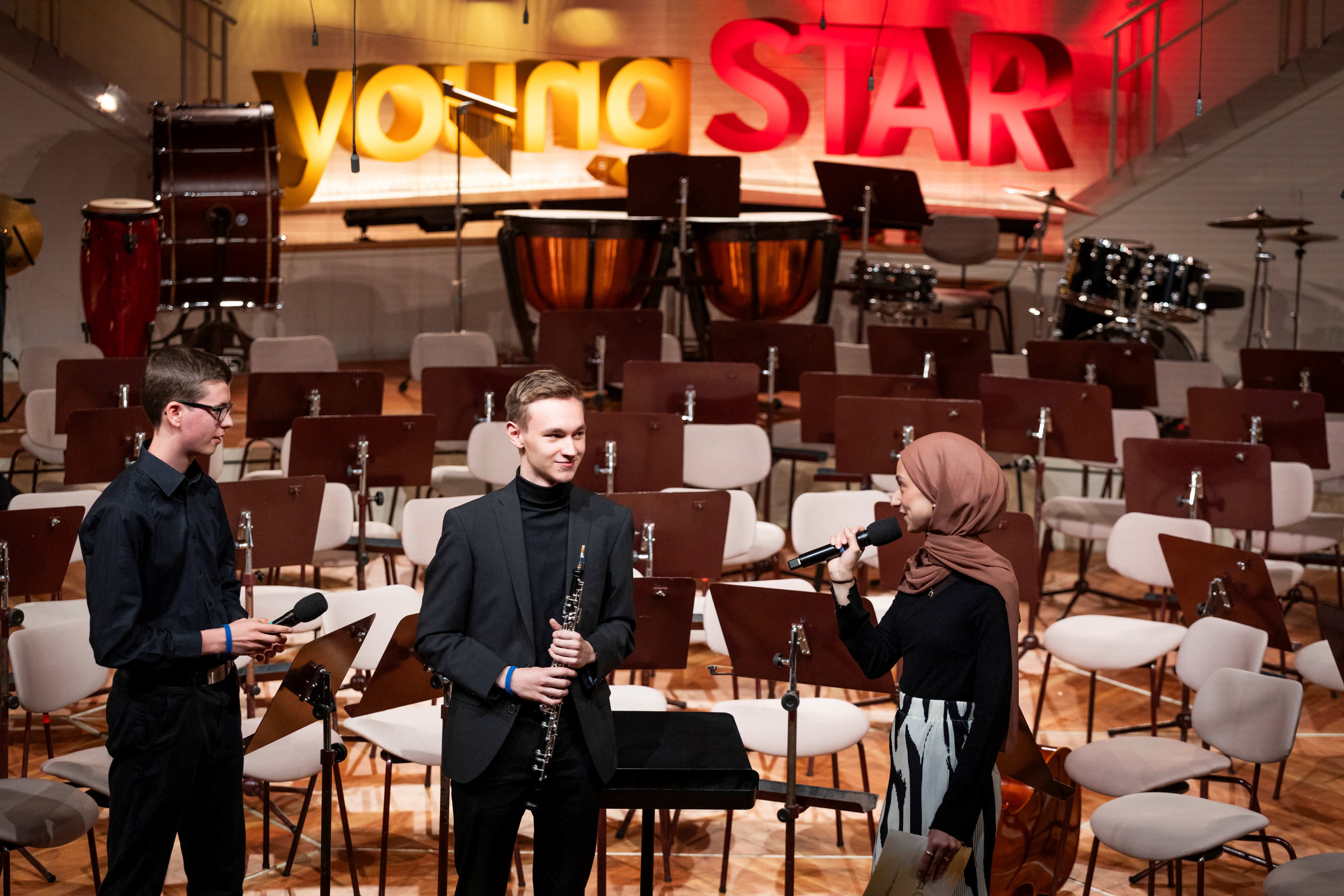Drei junge Leute stehen mit Mikrofonen und einer Klarinette auf der Bühne, vor leeren Notenständern und Schlaginstrumenten. Hinter ihnen leuchtet ein großes young STAR-Schild.