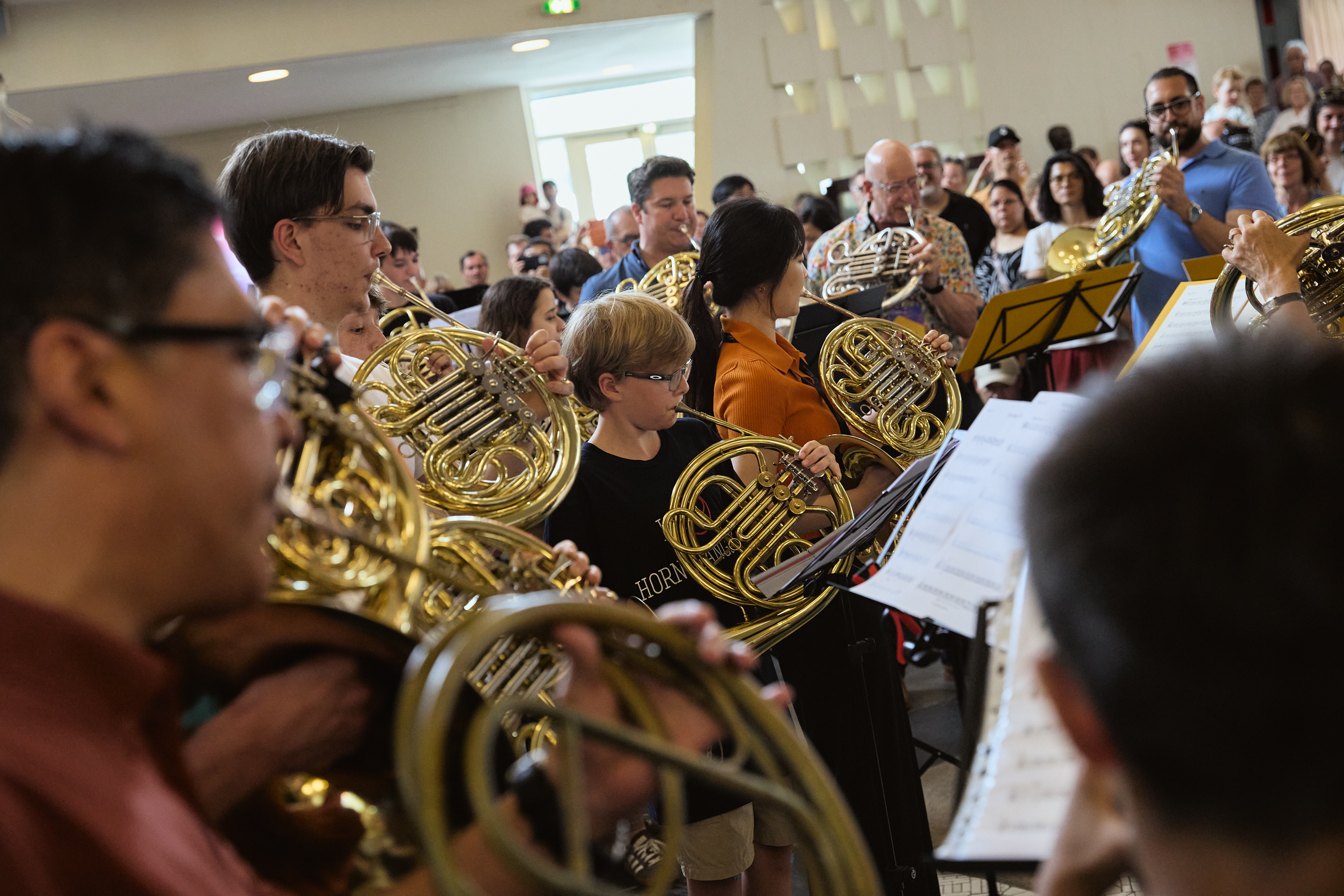 Eine Gruppe von Musikern, darunter Erwachsene und Kinder, spielt Waldhörner in einem überfüllten Innenraum, während im Hintergrund Zuschauer zuschauen.