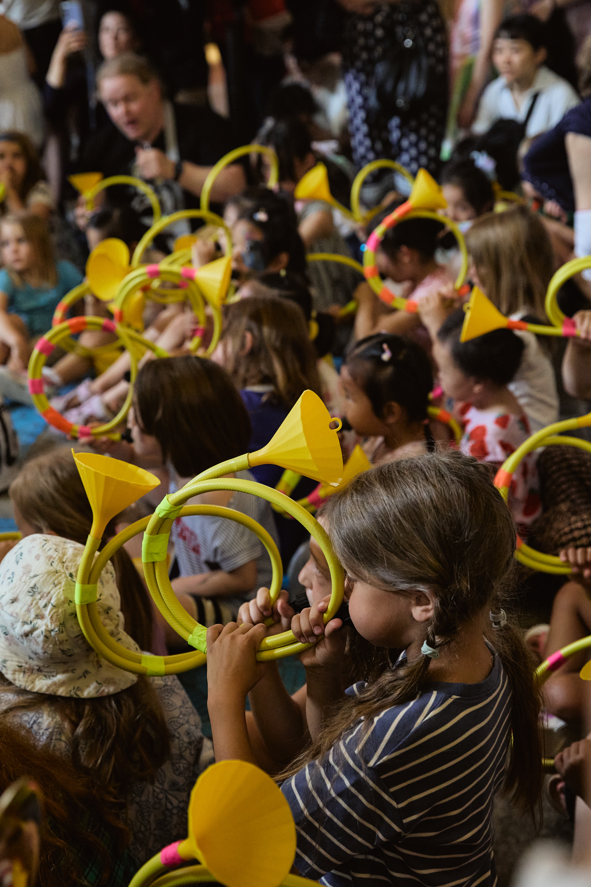 Die Kinder sitzen dicht beieinander, jedes hält eine leuchtend gelbe Plastiktröte in der Hand und bläst hinein. Viele tragen Partyhütchen aus Papier, und die Menge wirkt festlich und ist ganz in eine lebhafte Veranstaltung in den Innenräumen vertieft.