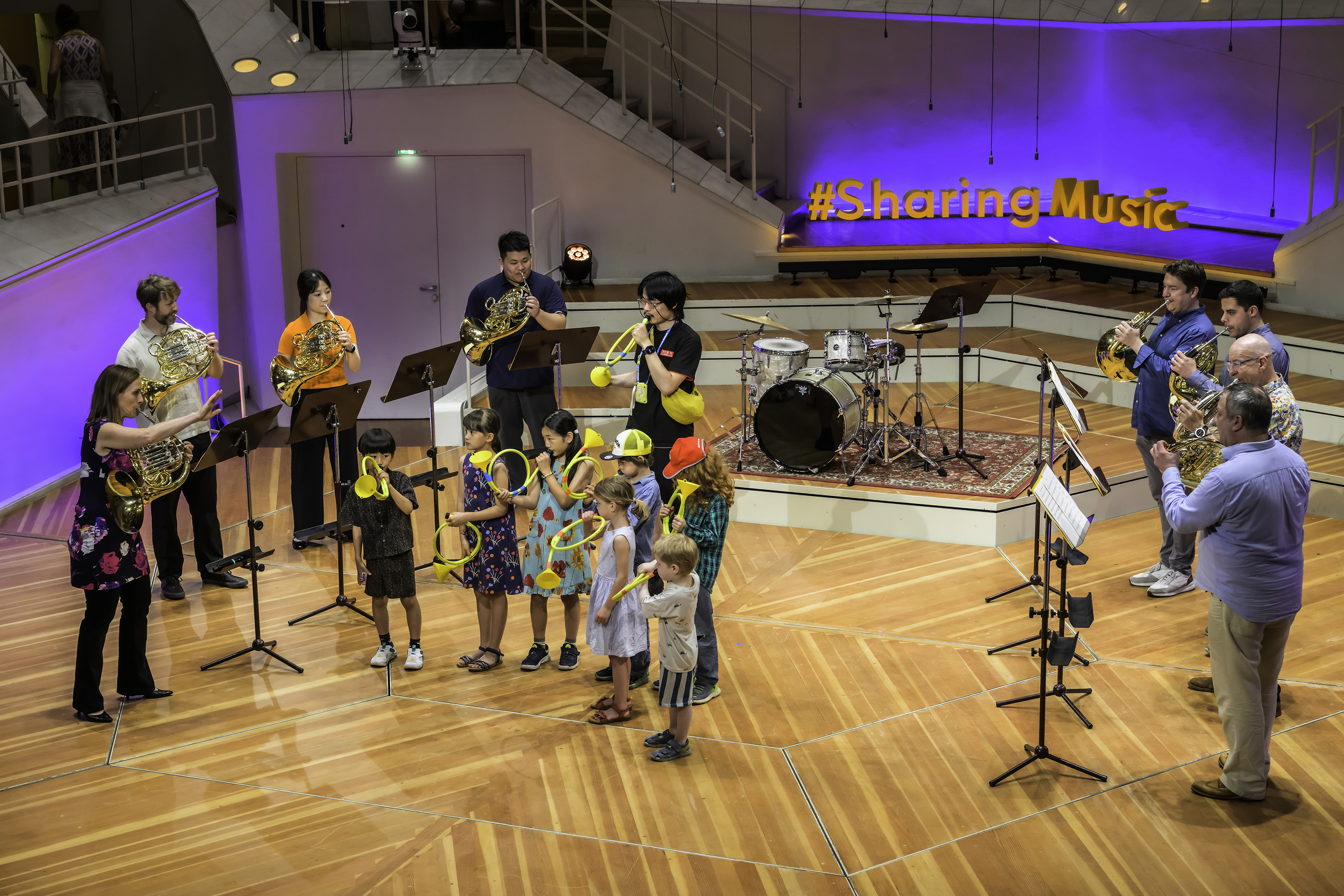 Eine Gruppe von Musikern und Kindern spielt mit verschiedenen Instrumenten auf einer Holzbühne, während im Hintergrund ein Schild mit der Aufschrift #SharingMusic leuchtet.