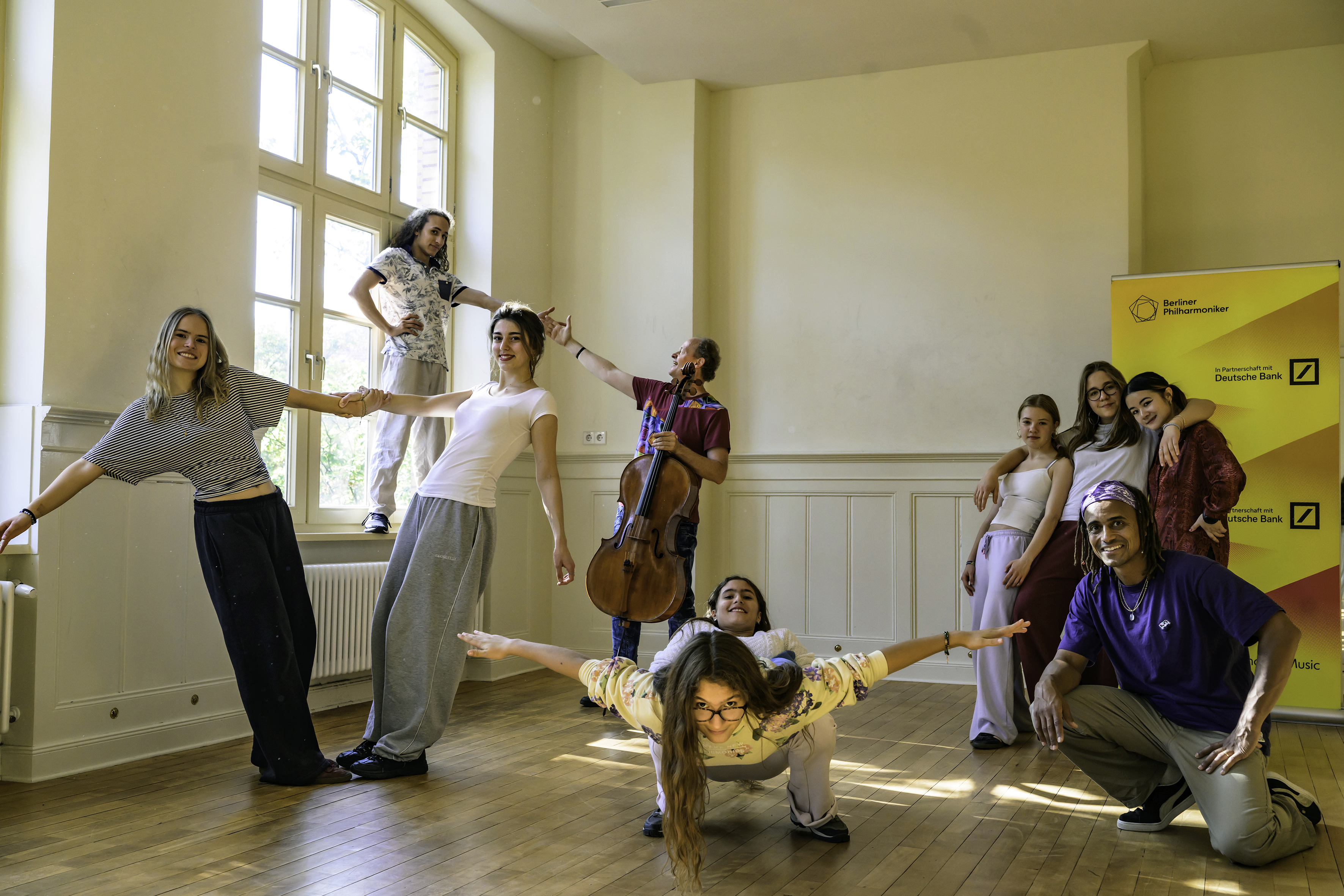 Eine Gruppe junger Menschen, die in einem Raum mit einem Cello kreativ posieren; im Vordergrund hockt eine Person, im Hintergrund ist ein Banner der Berliner Philharmoniker zu sehen.