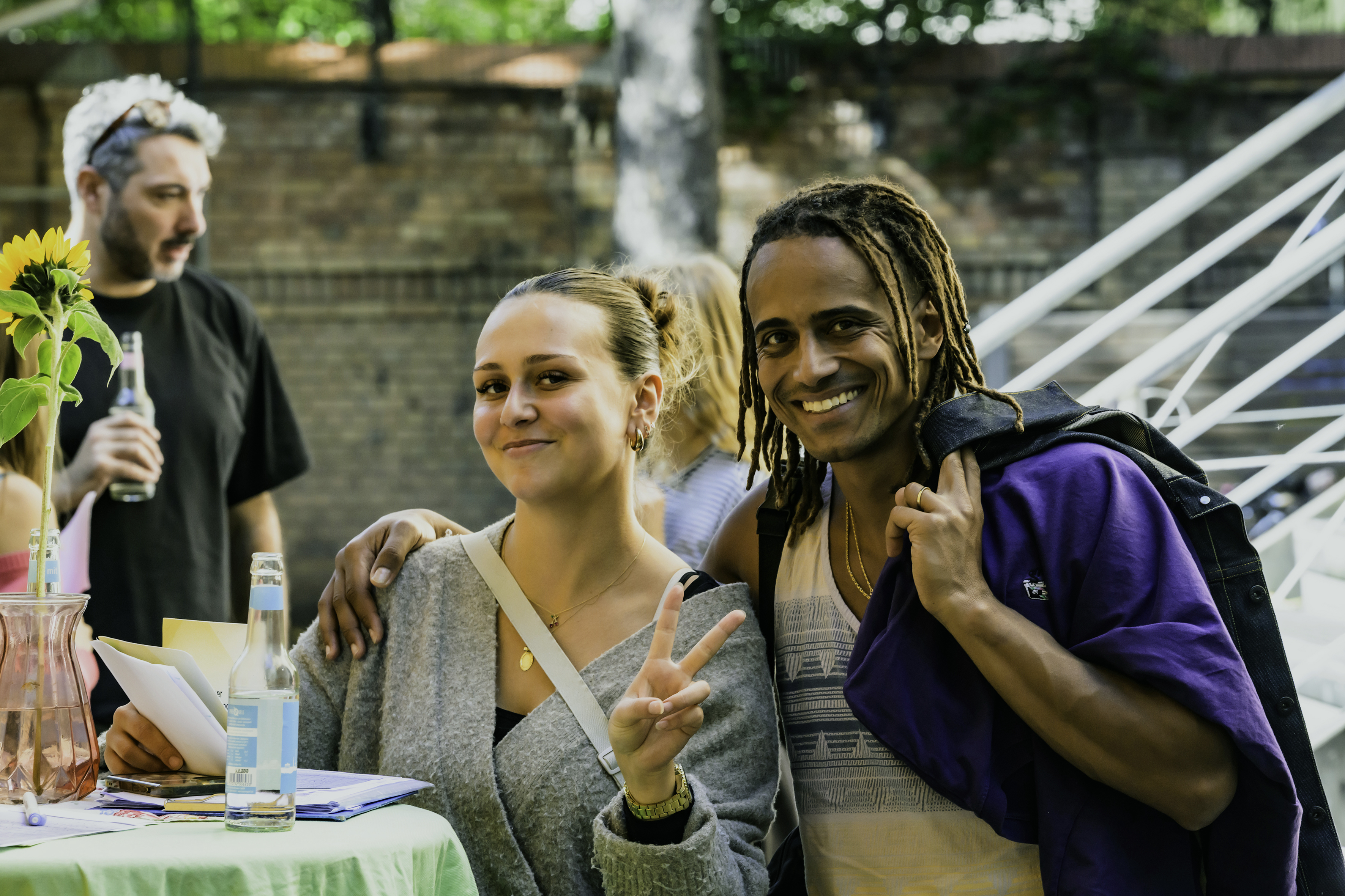 Eine lächelnde Frau, die das Peace-Zeichen macht, und ein Mann mit Dreadlocks, die gemeinsam draußen an einem Tisch stehen.