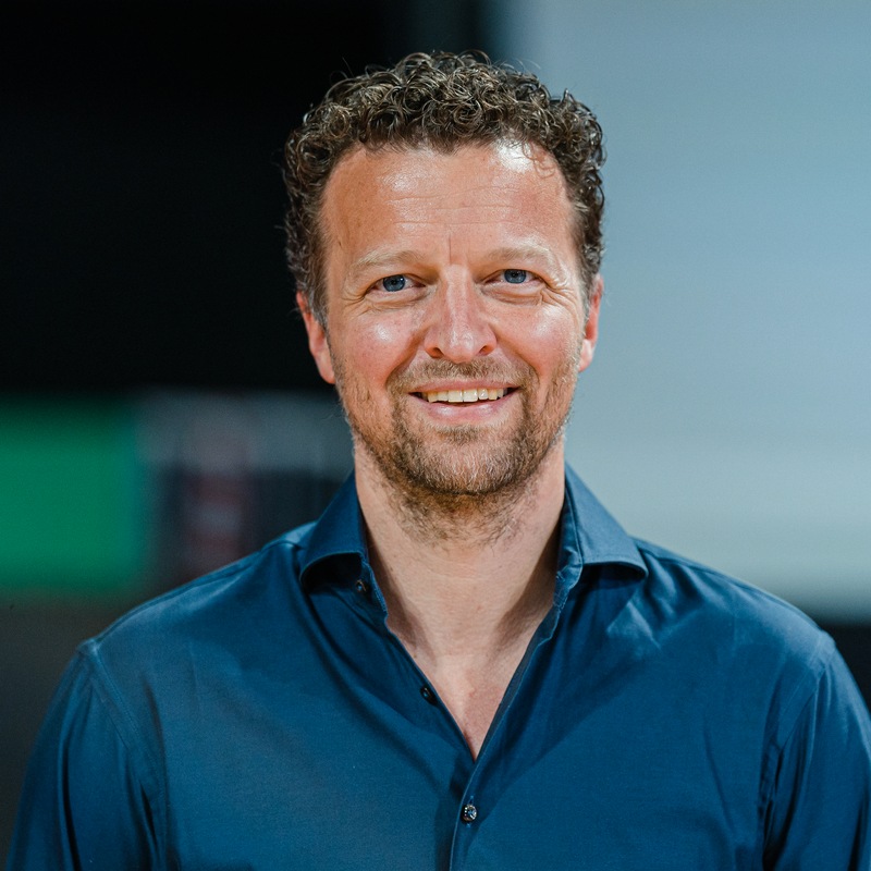 A man with curly brown hair and a beard, wearing a dark blue button-up shirt, smiles while standing indoors with a blurred background.