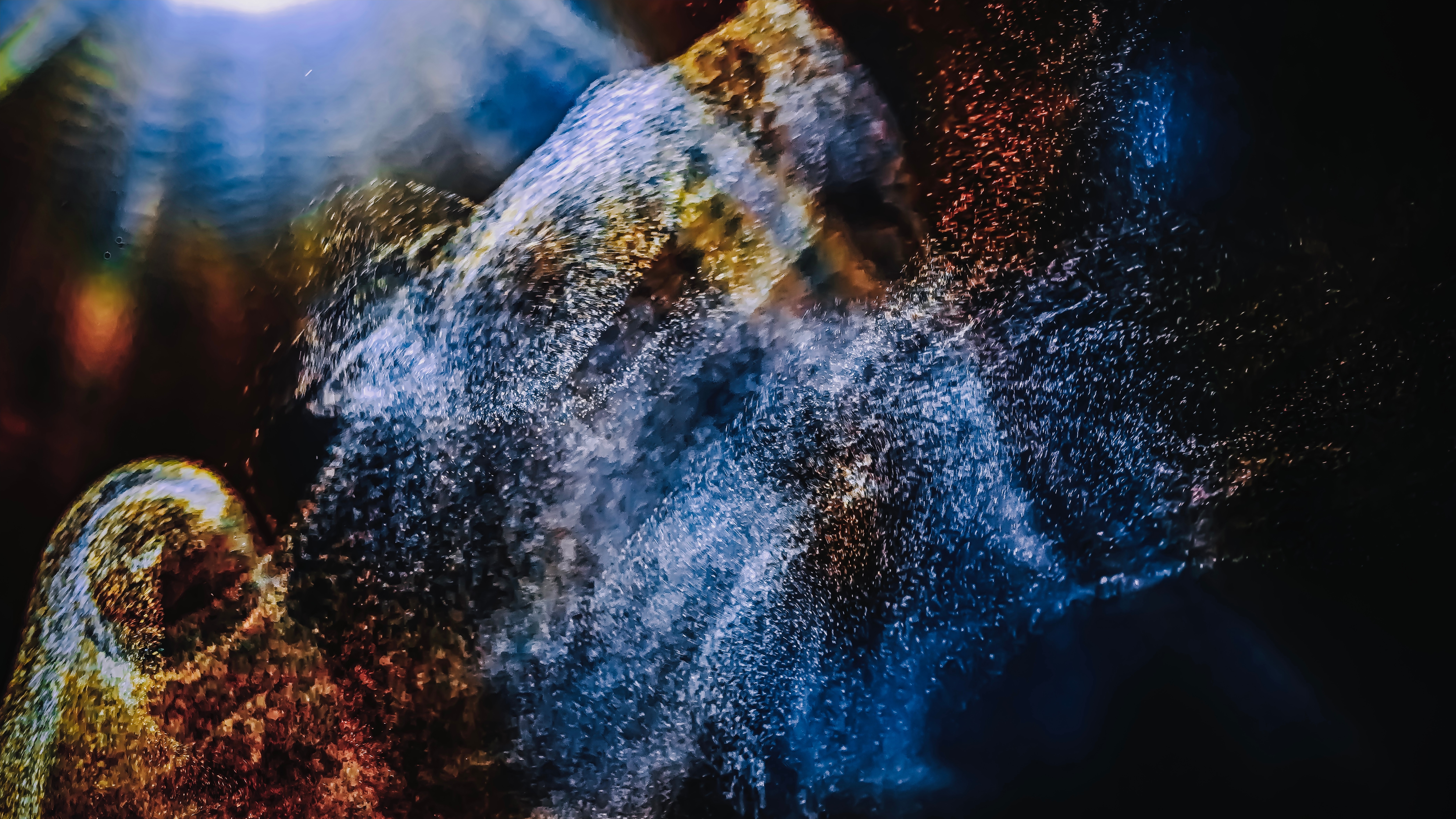 Abstraktes Bild von wirbelnden blauen, goldenen und roten Partikeln, die sich vor einem dunklen Hintergrund vermischen und einen dynamischen und lebendigen Effekt erzeugen, der an kosmischen Staub oder Lichtmuster unter Wasser erinnert.