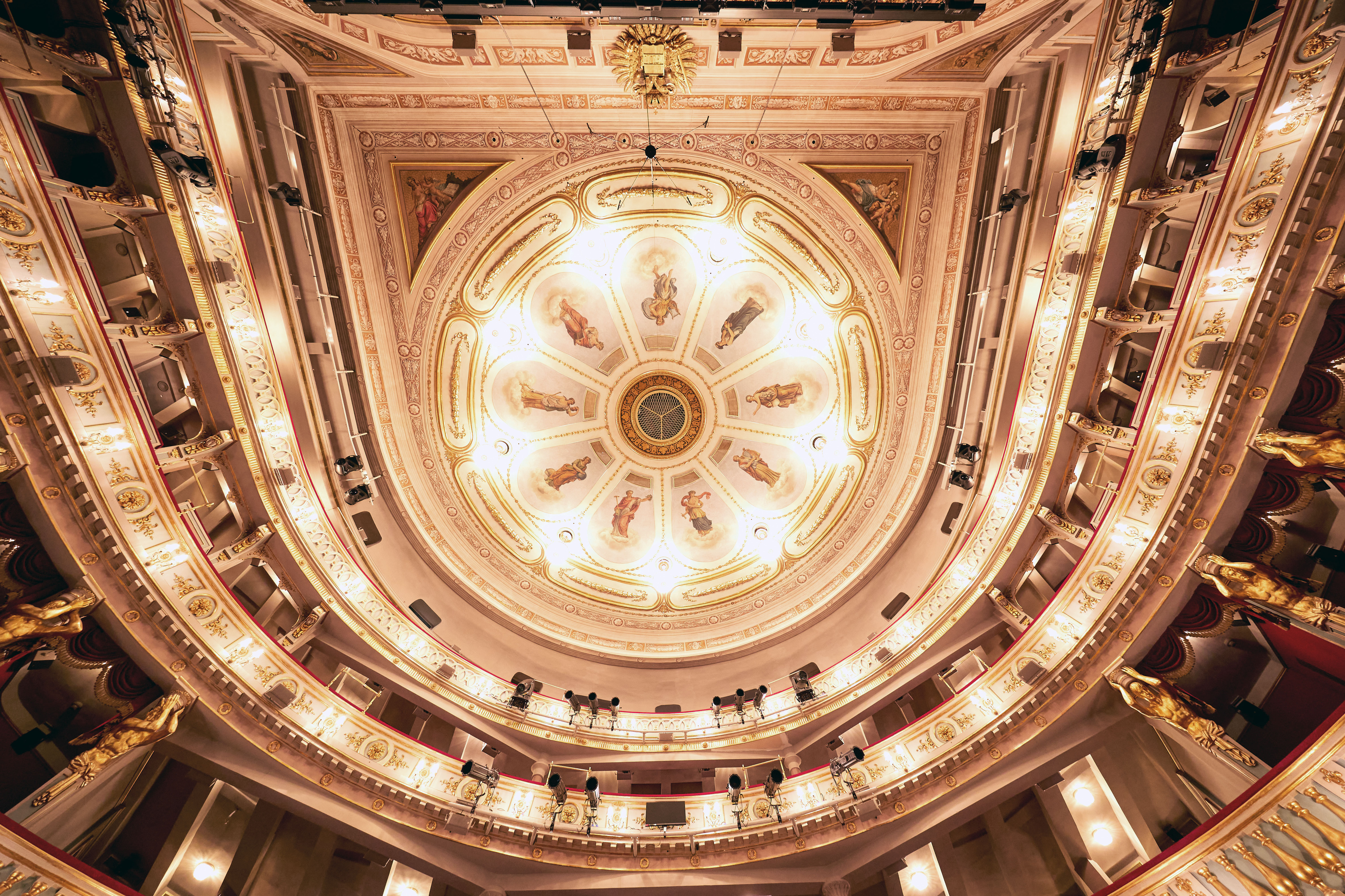 Verzierte, beleuchtete Decke des Theater an der Wien mit aufwendigen Dekorationen, bemalten Tafeln, goldenen Akzenten und um den Innenraum geschwungenen Balkonen, von unten betrachtet.