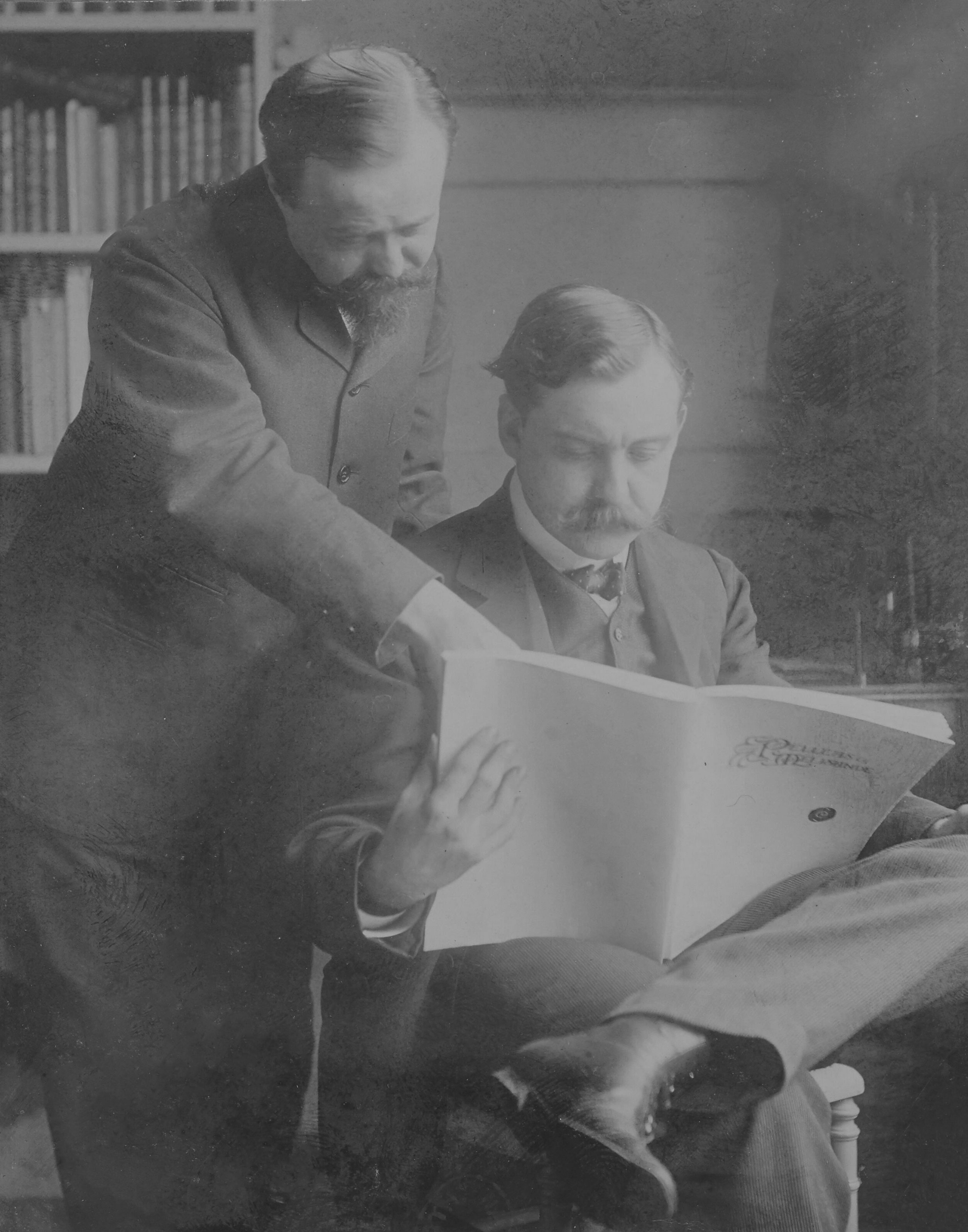 Two men in suits with mustaches look at a large open book; one is seated holding the book, the other stands beside him, pointing at a page. Bookshelves are visible in the background.