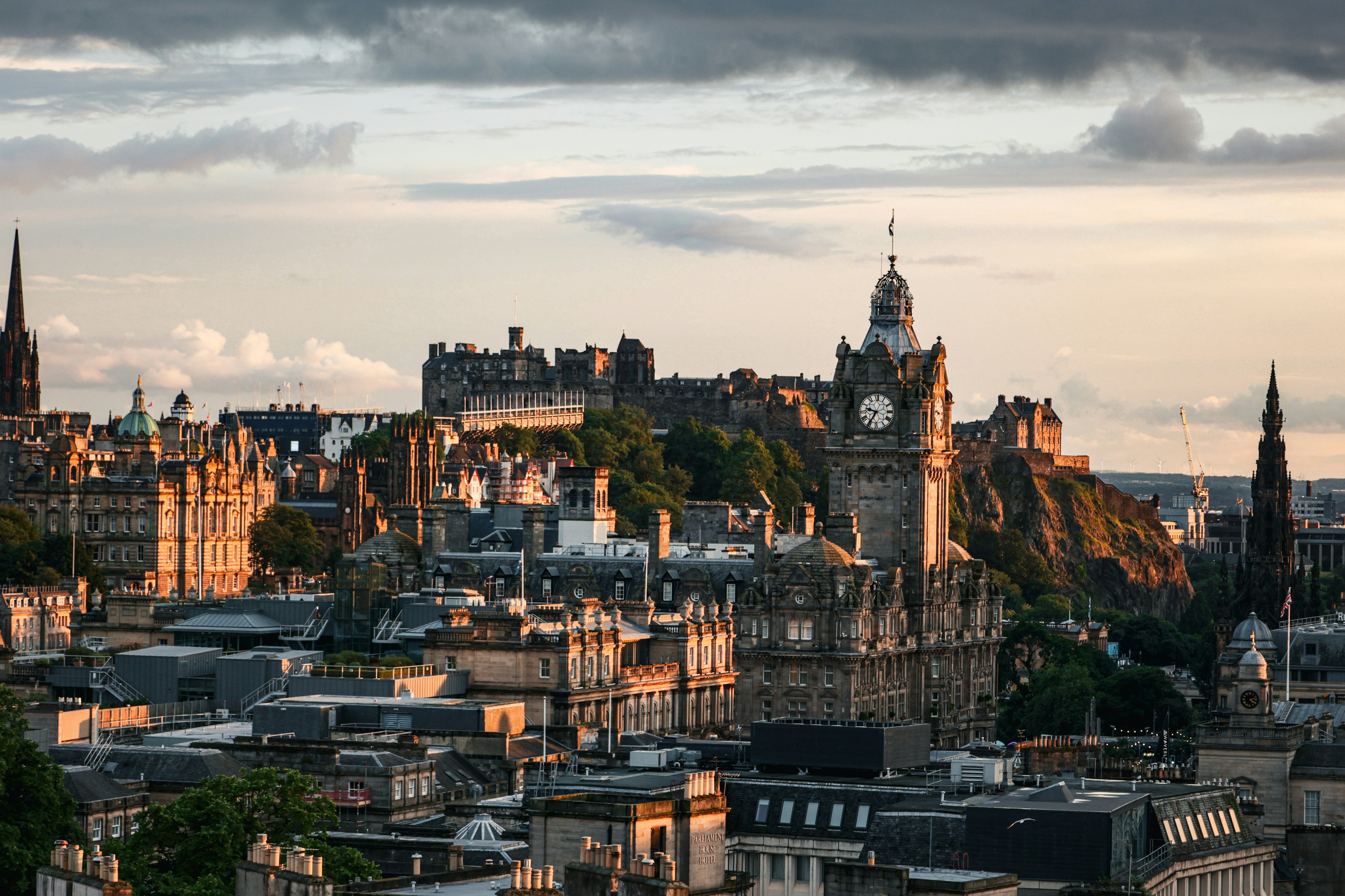 Edinburgh cityscape at sunset, featuring the historic Edinburgh Castle on a hill, the Balmoral Hotel clock tower, and various old stone buildings under a partly cloudy sky.