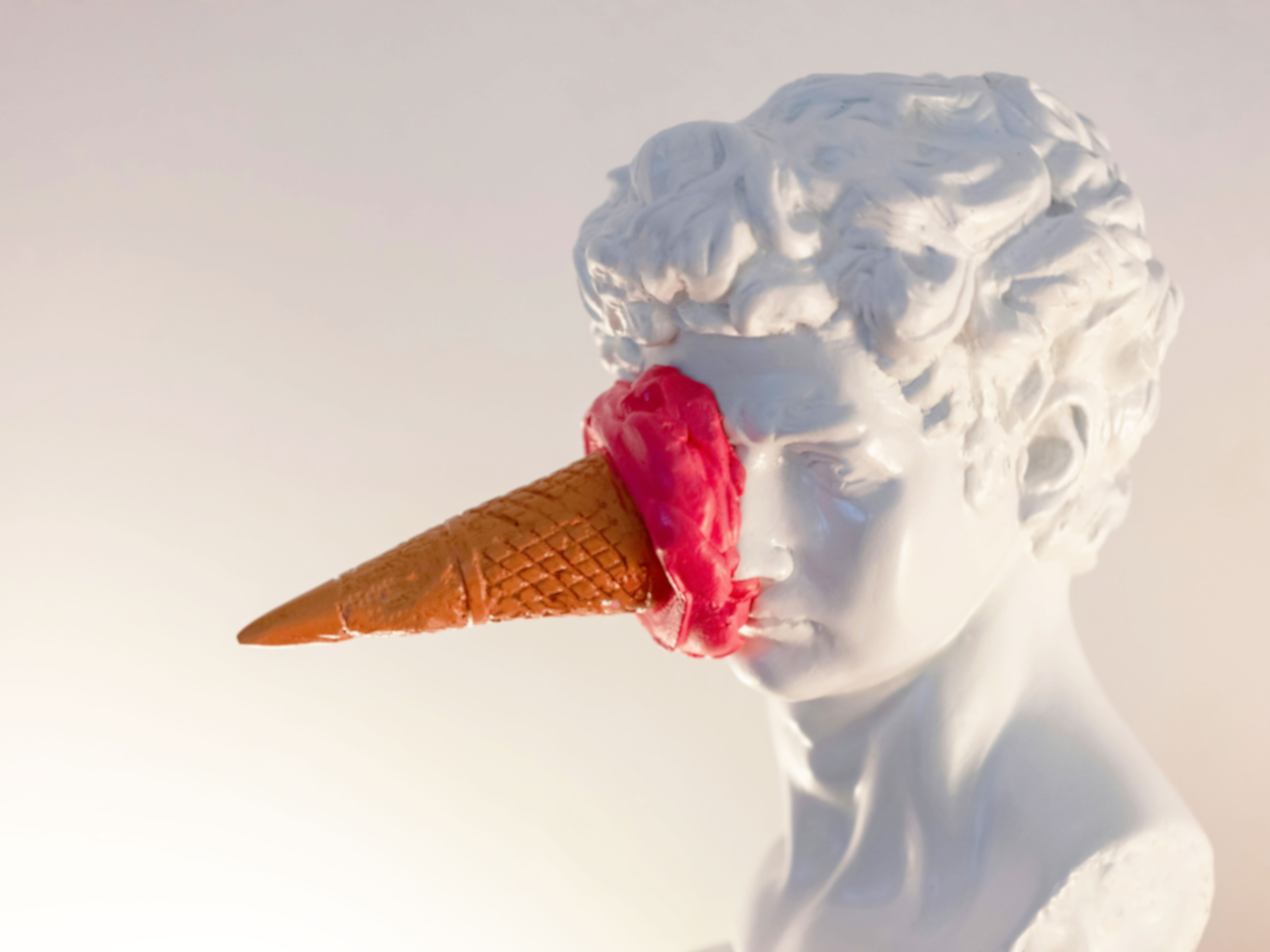 Eine weiße Büste, auf deren Nase ein orangefarbenes Eiswaffelhörnchen steckt, umgeben von schmelzendem rosa Eis.