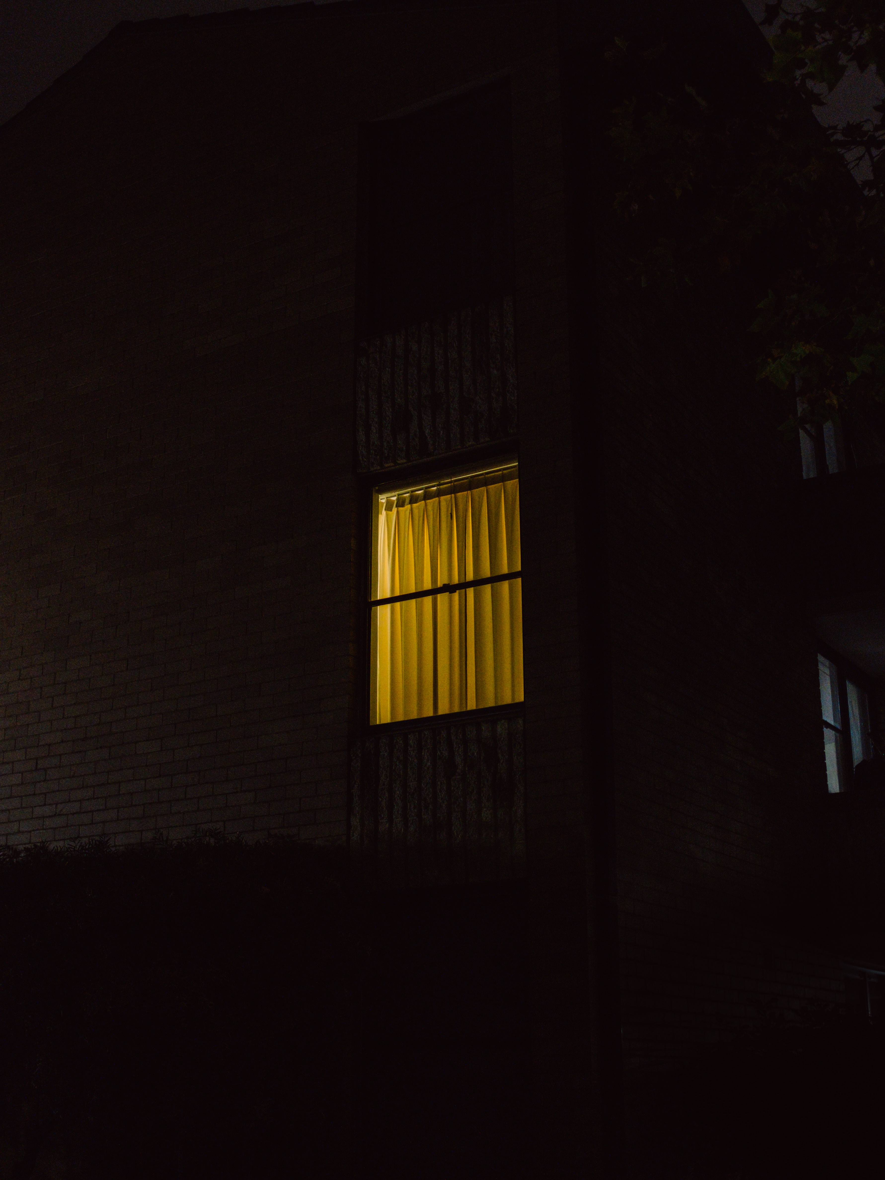 A dark building at night with one window illuminated by warm yellow light behind closed curtains, creating a sense of mystery and contrast against the surrounding darkness.